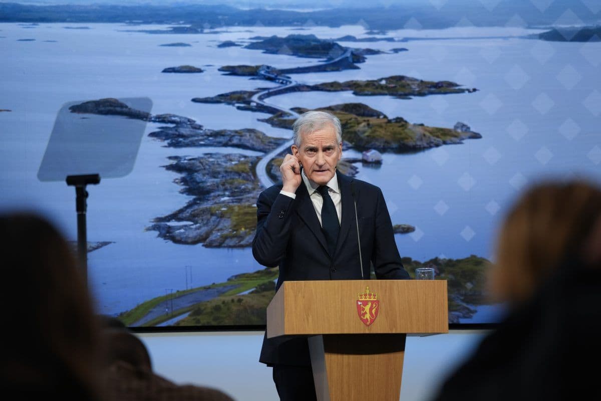 En mann i dress står på et podium med det norske riksvåpenet og taler foran en skjerm som viser et kystlandskap med svingete veier.
