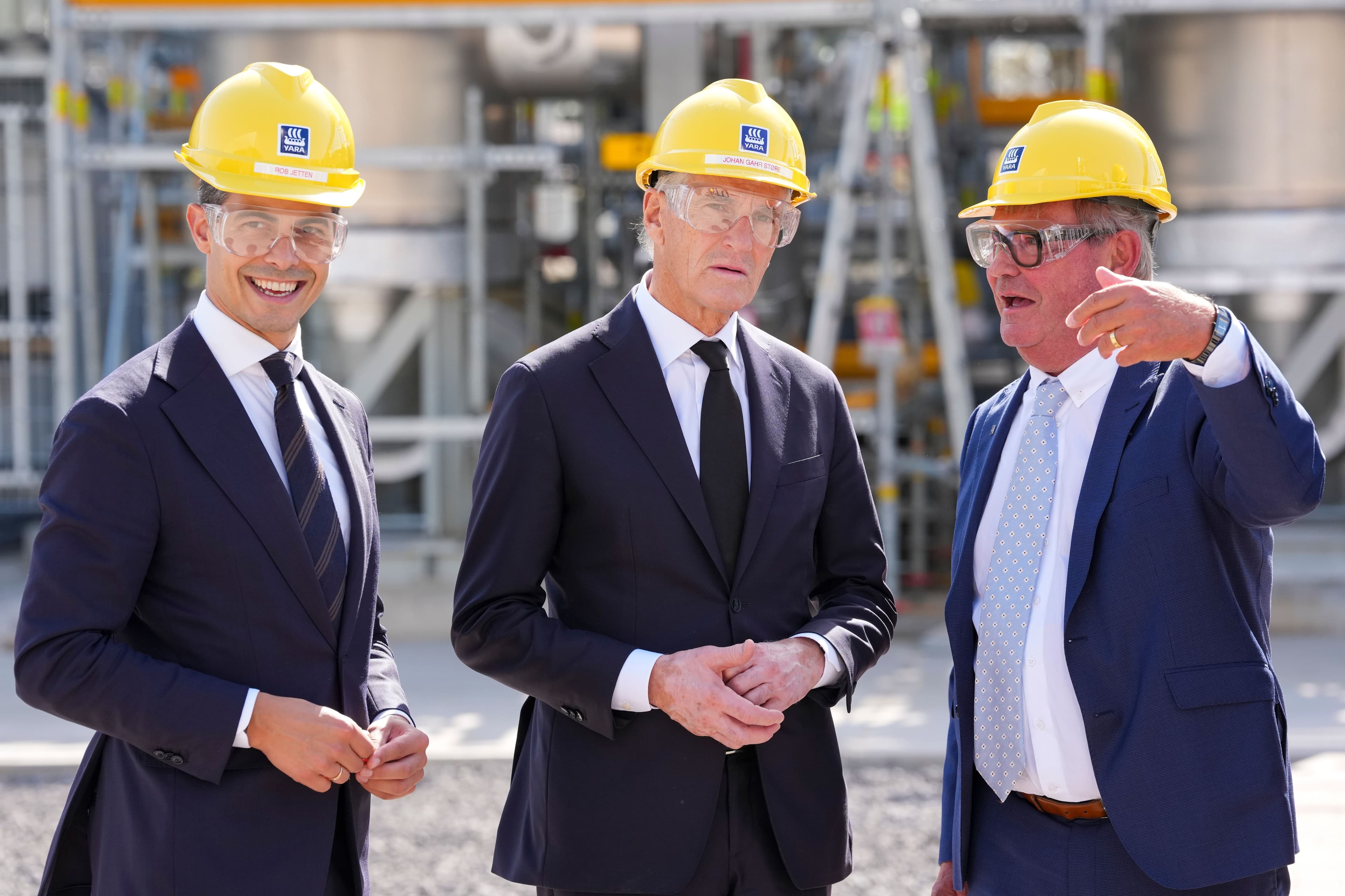 Tre menn i dress, gule vernehjelmer og vernebriller står på et industrianlegg.