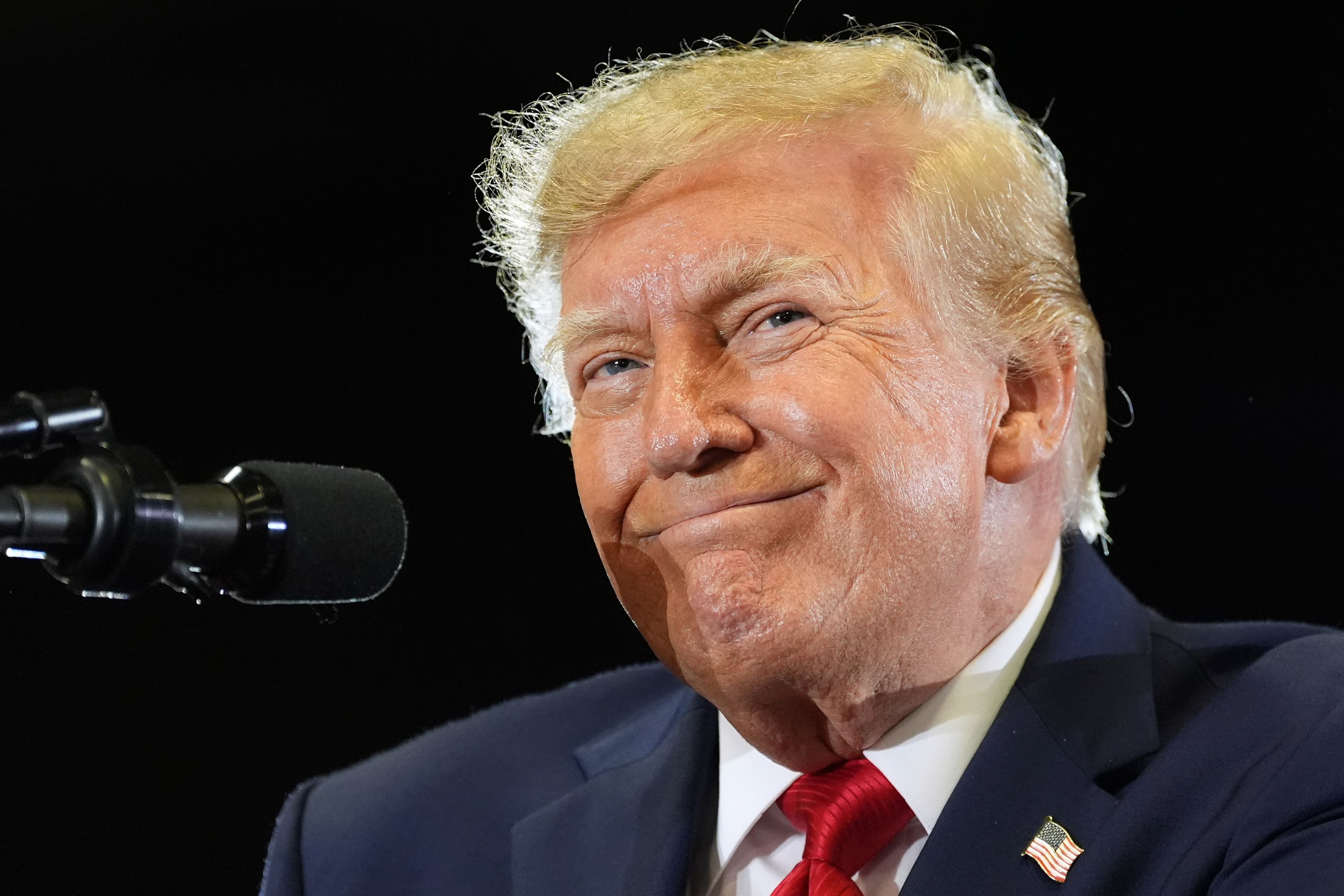 Donald Trump smiler, iført en blå dress, rødt slips og en jakkeslagsnål med det amerikanske flagget, med en mikrofon til venstre for seg.