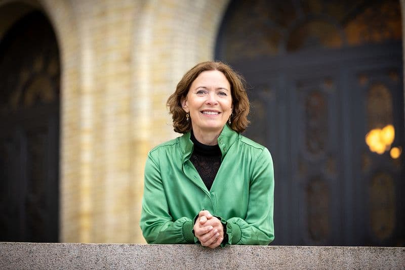 En smilende kvinne i grønn bluse lener seg mot et rekkverk av stein, med en bygning med buet inngang bak henne.