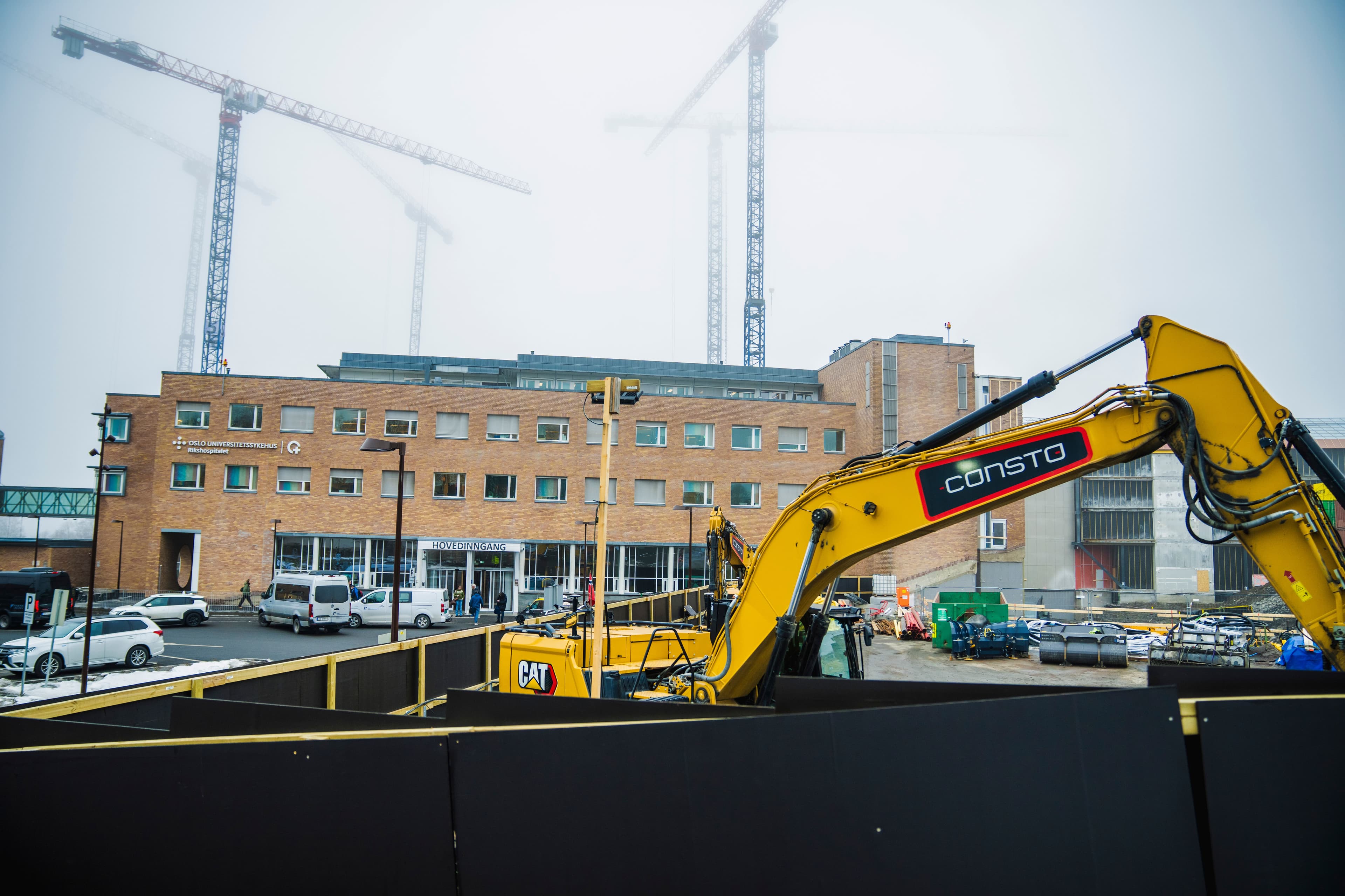 En gul «CONSTO»-gravemaskin på en byggeplass med en stor mursteinsbygning og flere kraner i en tåkete bakgrunn.