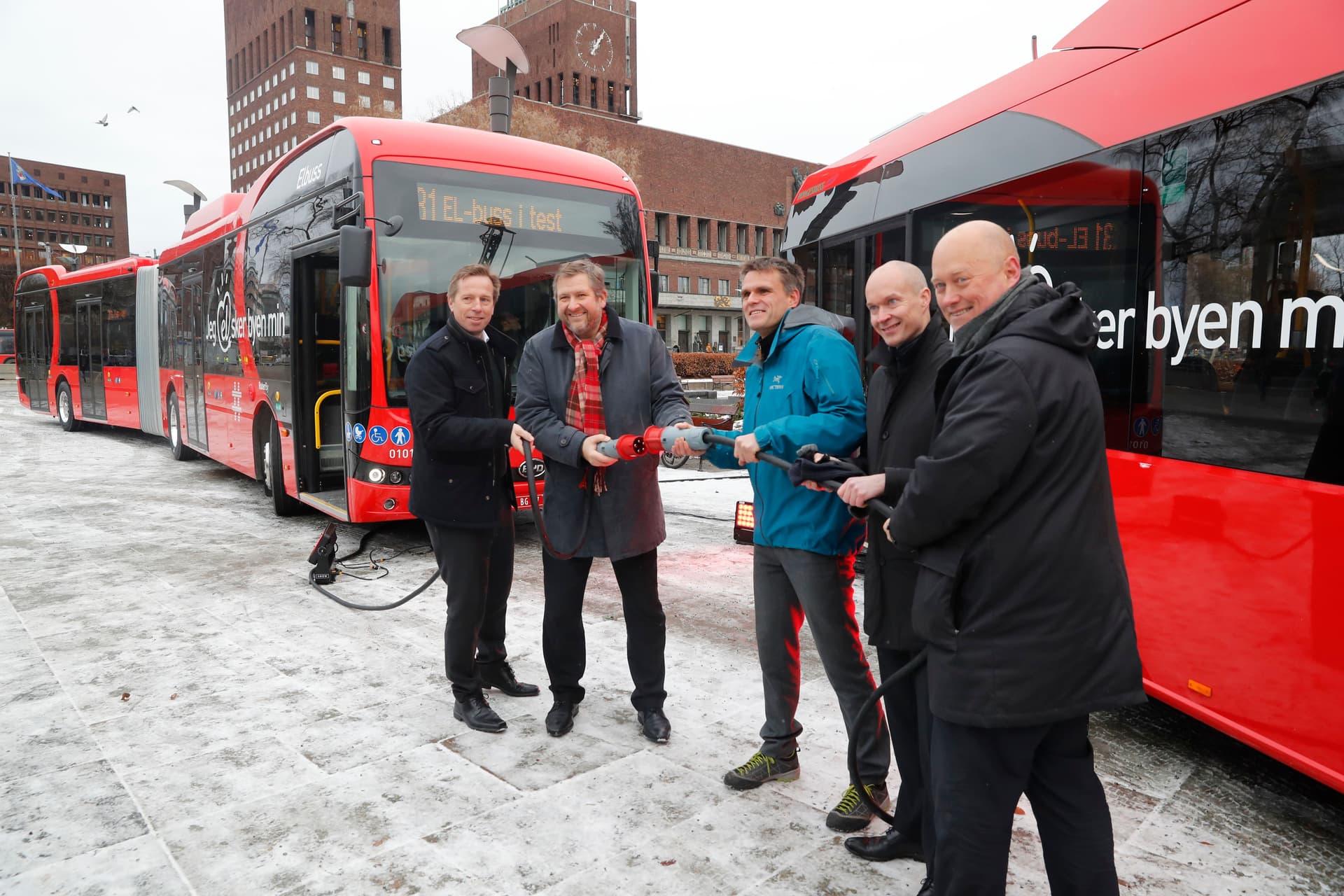 Fem menn holder en ladekabel foran to røde elektriske busser, med en bygning i byen i den snødekte bakgrunnen.