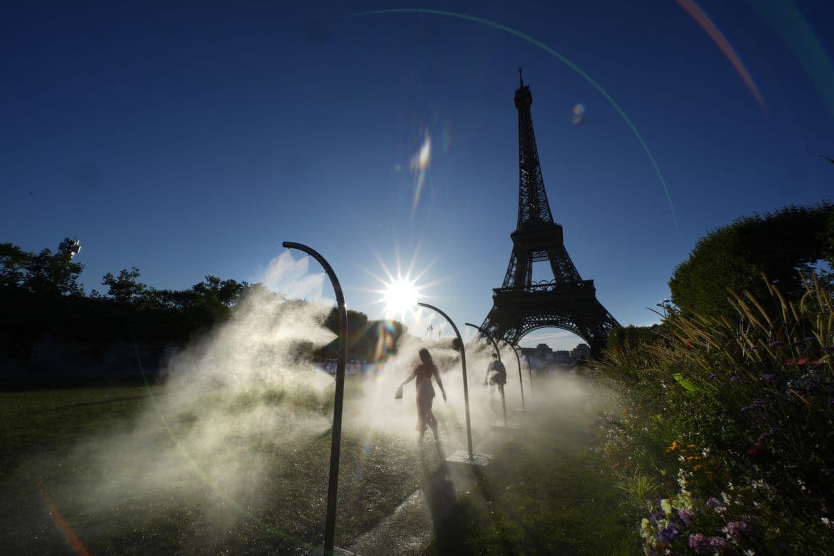 Folk går gjennom tåkebuer i nærheten av Eiffeltårnet i Paris, mens sollyset skinner bak og skaper silhuetter og reflekser.