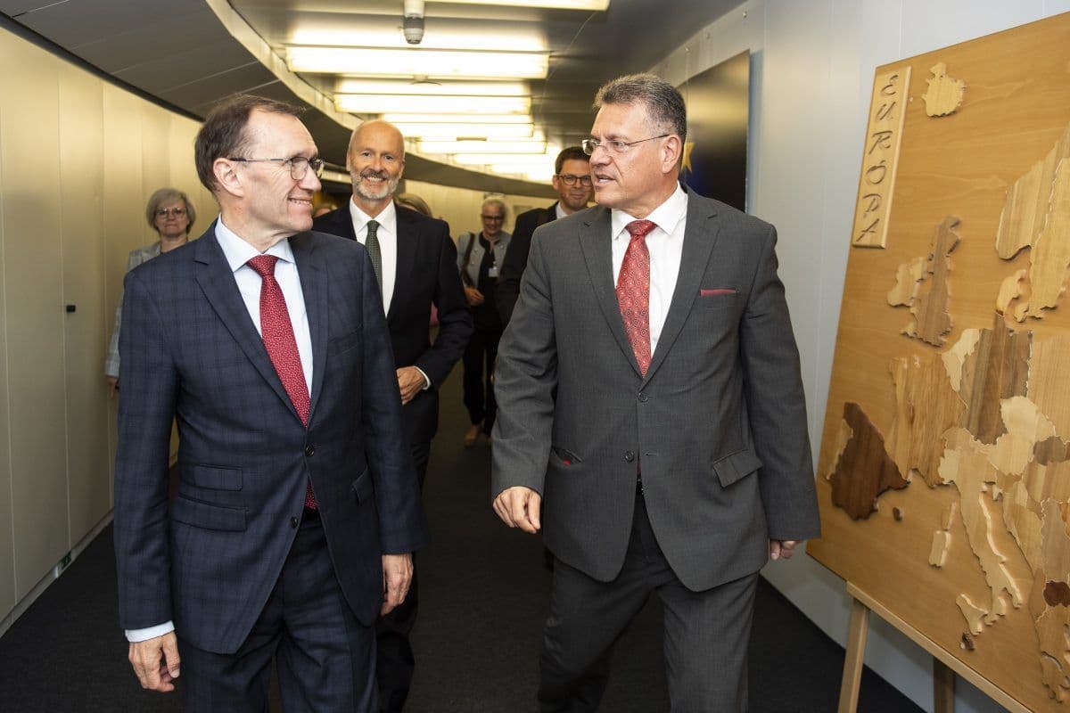 To menn i dress går og snakker i en gang. Et kart merket "Europa" er synlig på et stativ i nærheten. Andre personer i formell antrekk er i bakgrunnen.