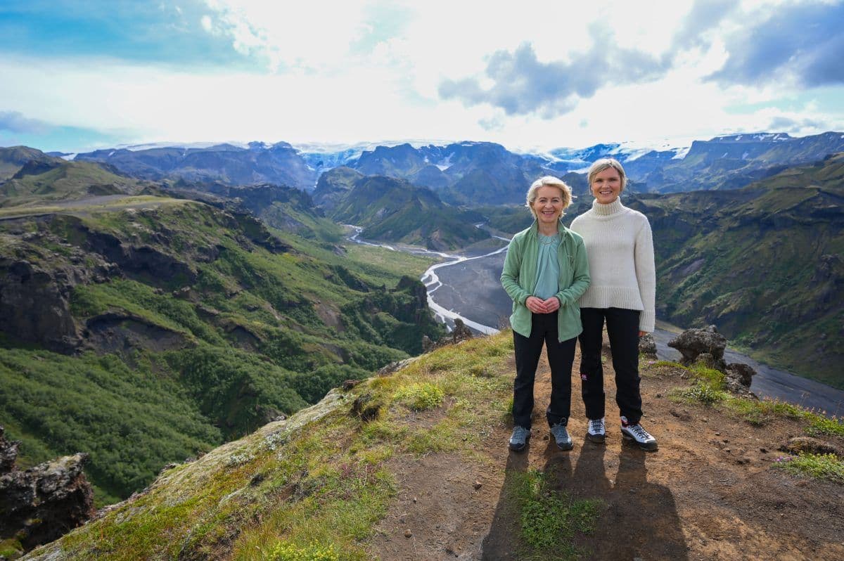 To kvinner står på en gresskledd klippe med utsikt over en dal med en svingete elv, fjell og en delvis overskyet himmel i bakgrunnen.