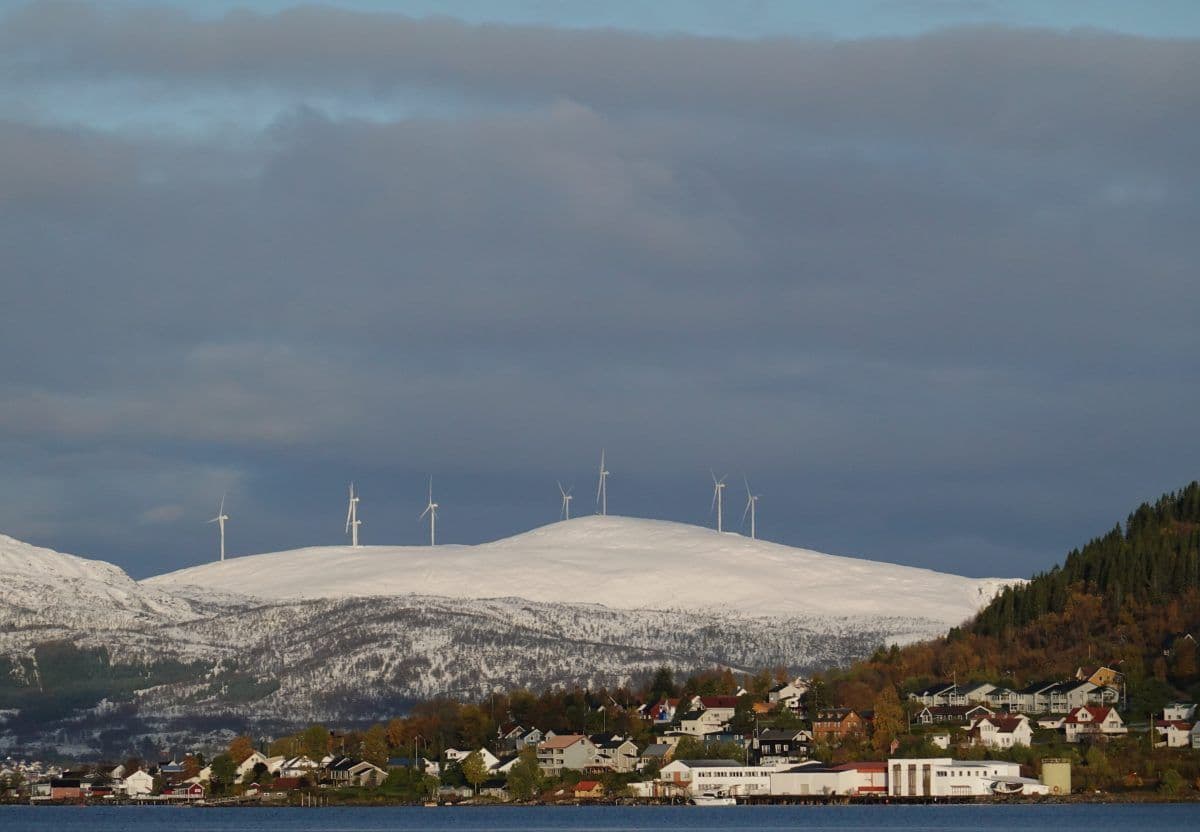 Kystby med hus i forgrunnen og flere vindturbiner på en snødekt høyde under overskyet himmel.
