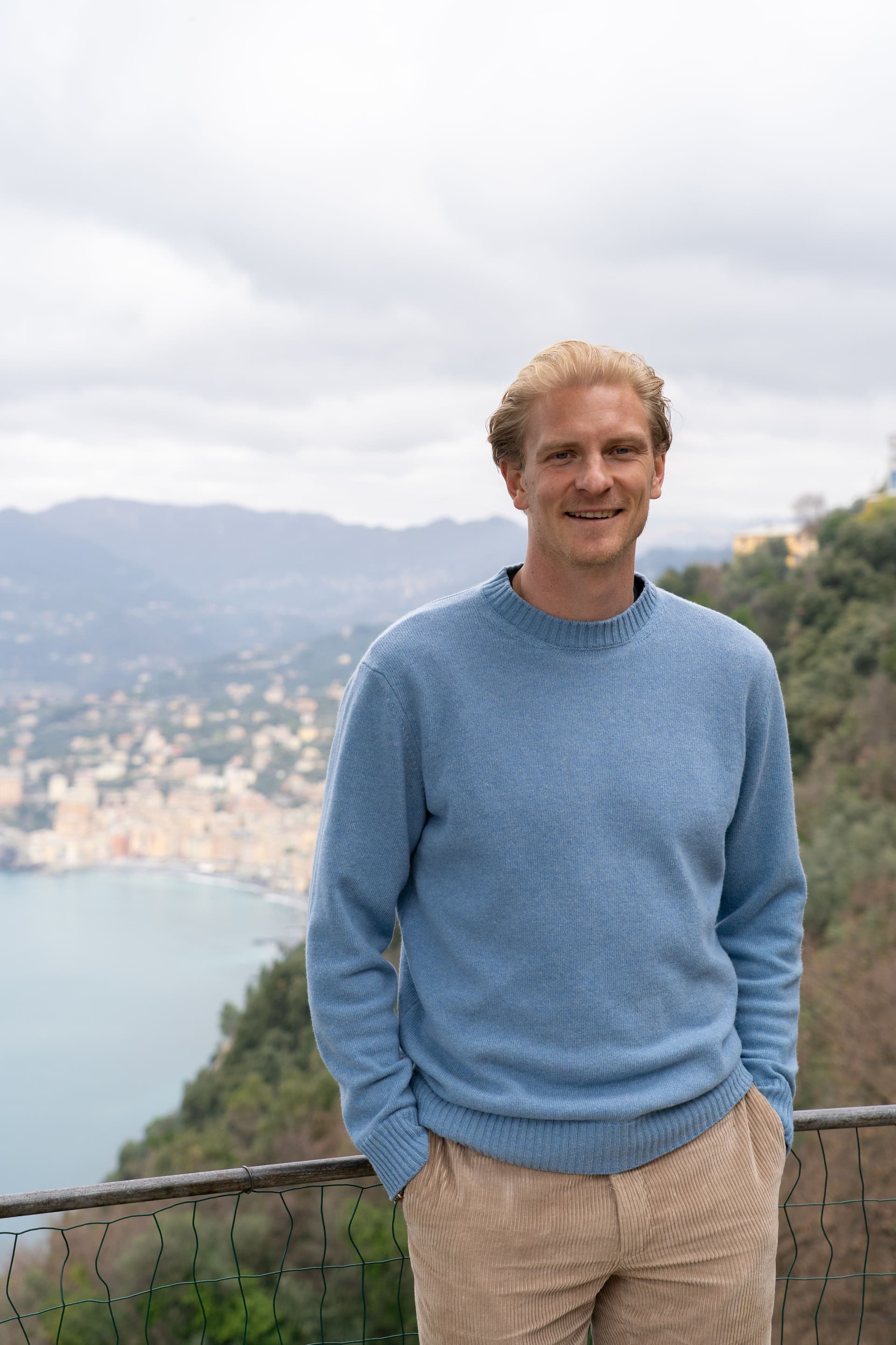 A smiling man in a light blue sweater and khaki pants stands on a hill overlooking a coastal town and bay.