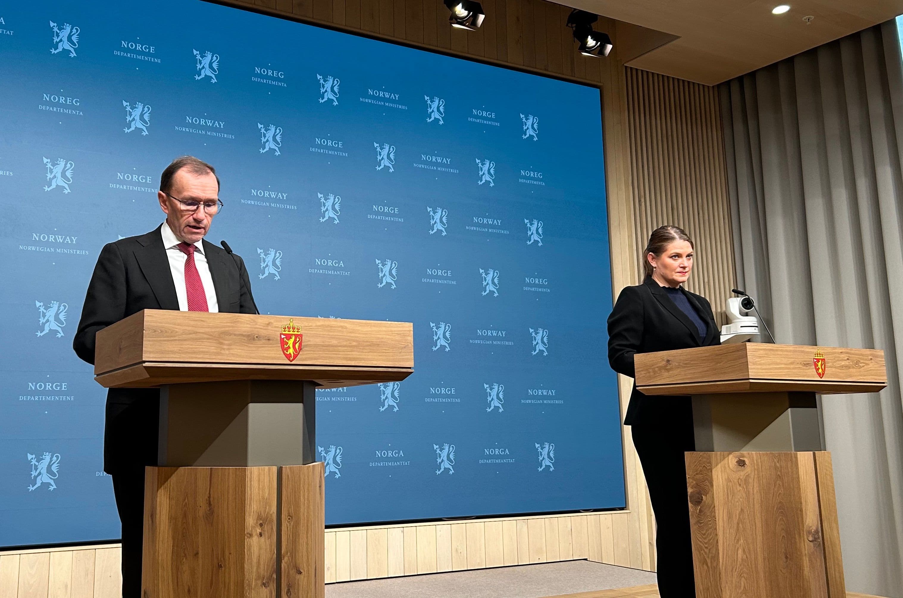 En mann taler fra en talerstol mens en kvinne står i nærheten, begge foran en bakgrunn med den norske regjeringens profilering.