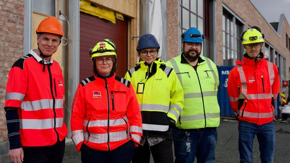 Fem personer iført refleksvester og hjelmer står utenfor en industribygning på en overskyet dag.