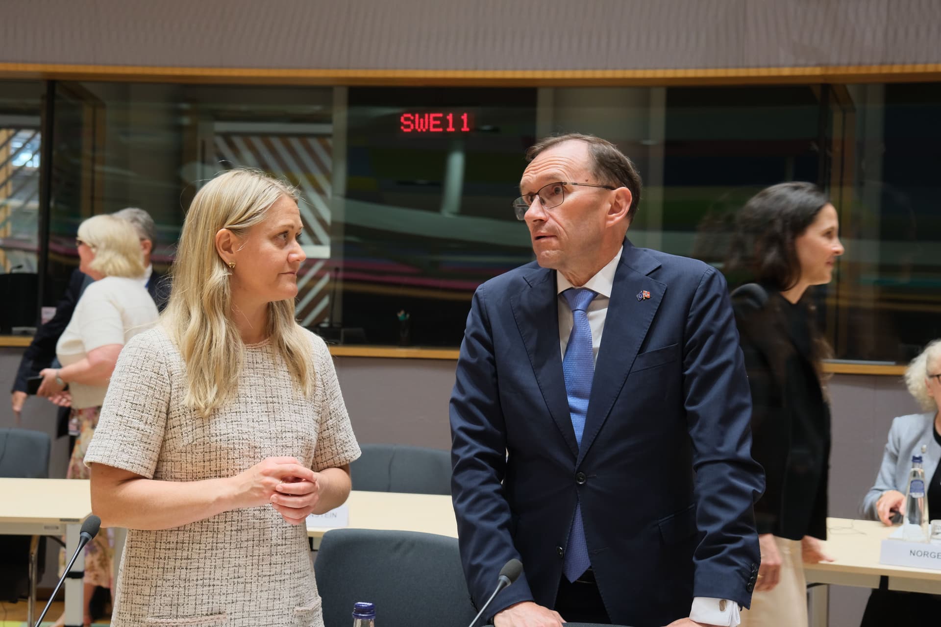 A woman in a light patterned dress and a man in a dark suit and glasses stand in a modern conference room.
