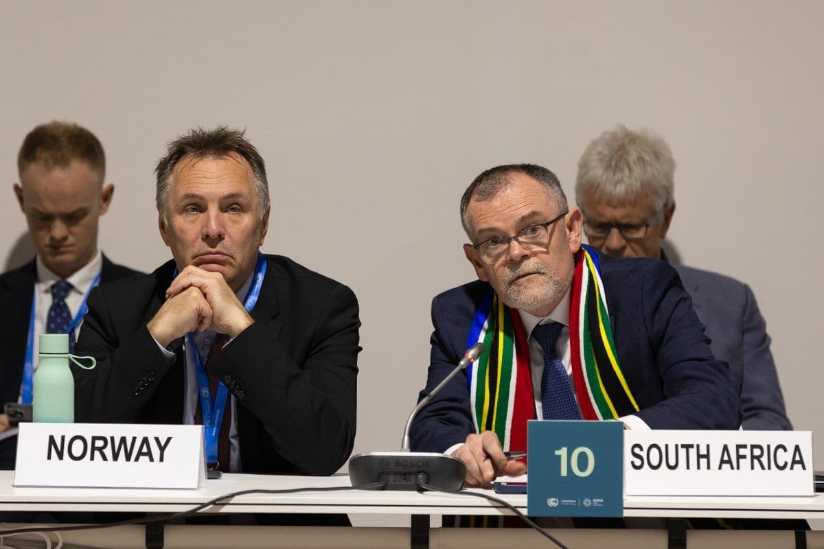 To menn i dress sitter ved et konferansebord. Den ene representerer Norge, den andre Sør-Afrika. De lytter oppmerksomt, med navneskilt og mikrofoner foran seg.