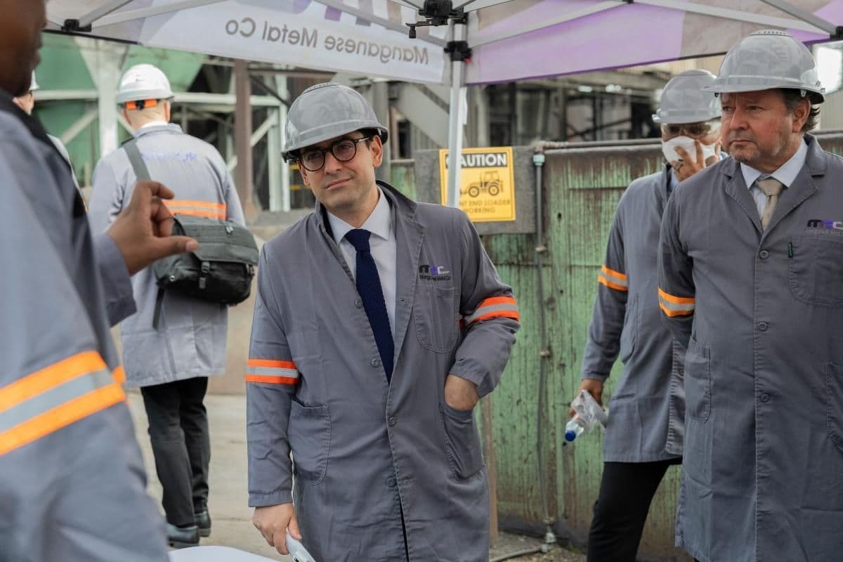 En gruppe mennesker i grå vernefrakker og hjelmer står under et telt på et industriområde, med sikkerhetsskilt synlige i bakgrunnen.