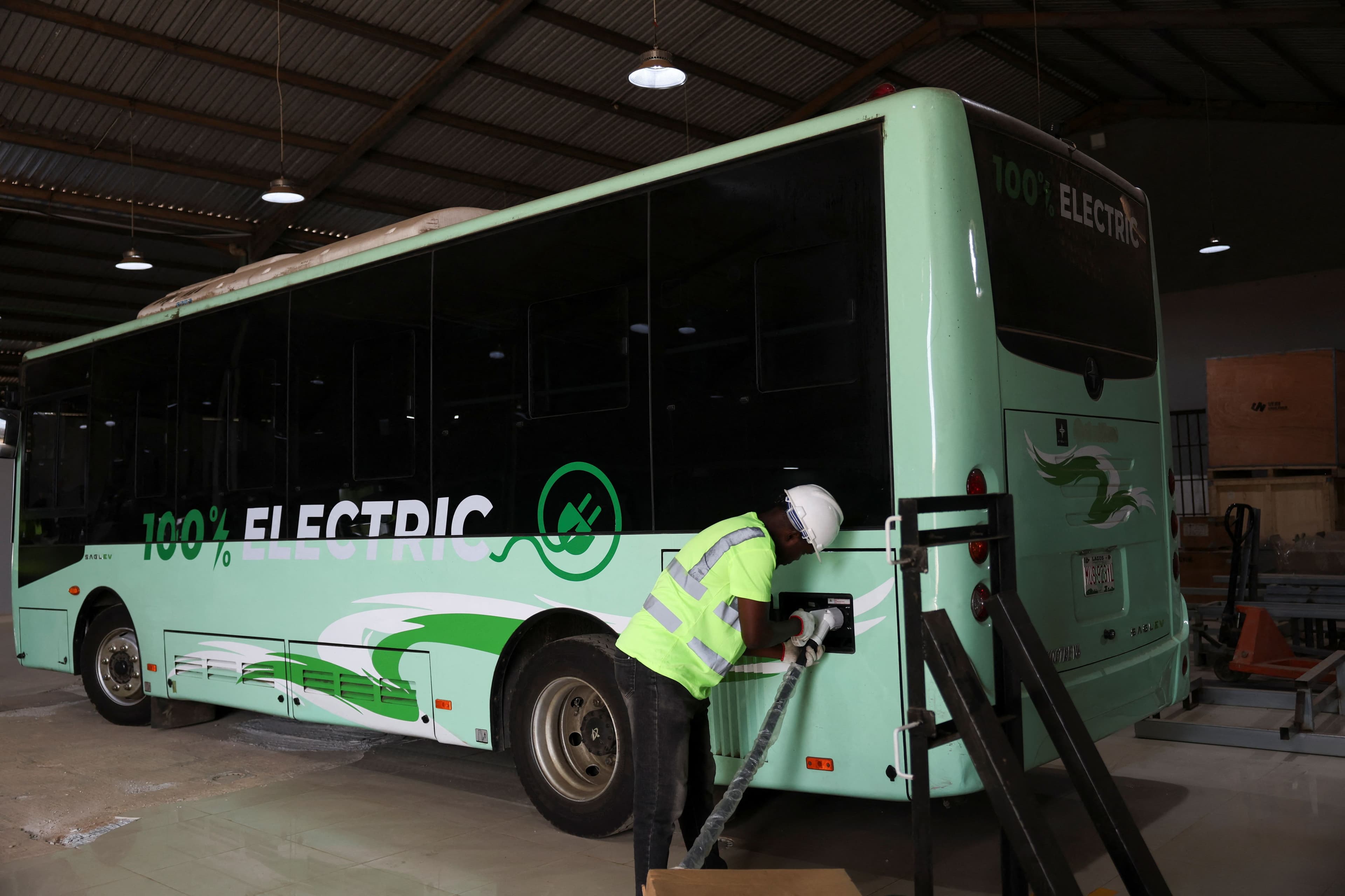 En arbeider lader en lysegrønn elektrisk buss med påskriften «100 % ELEKTRISK» i et innendørsanlegg.