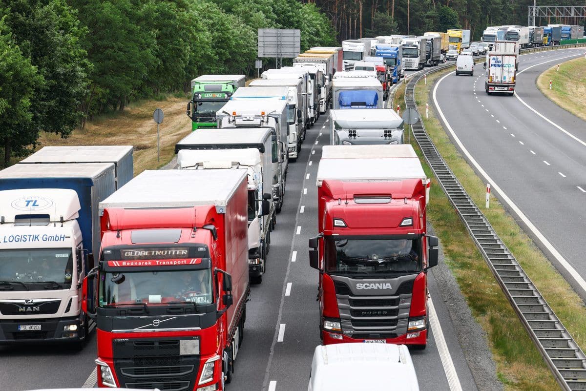 En rekke lastebiler står i kø på en flerfelts motorvei, med flere kjøretøy synlige i begge retninger. Trær står langs veien på den ene siden.