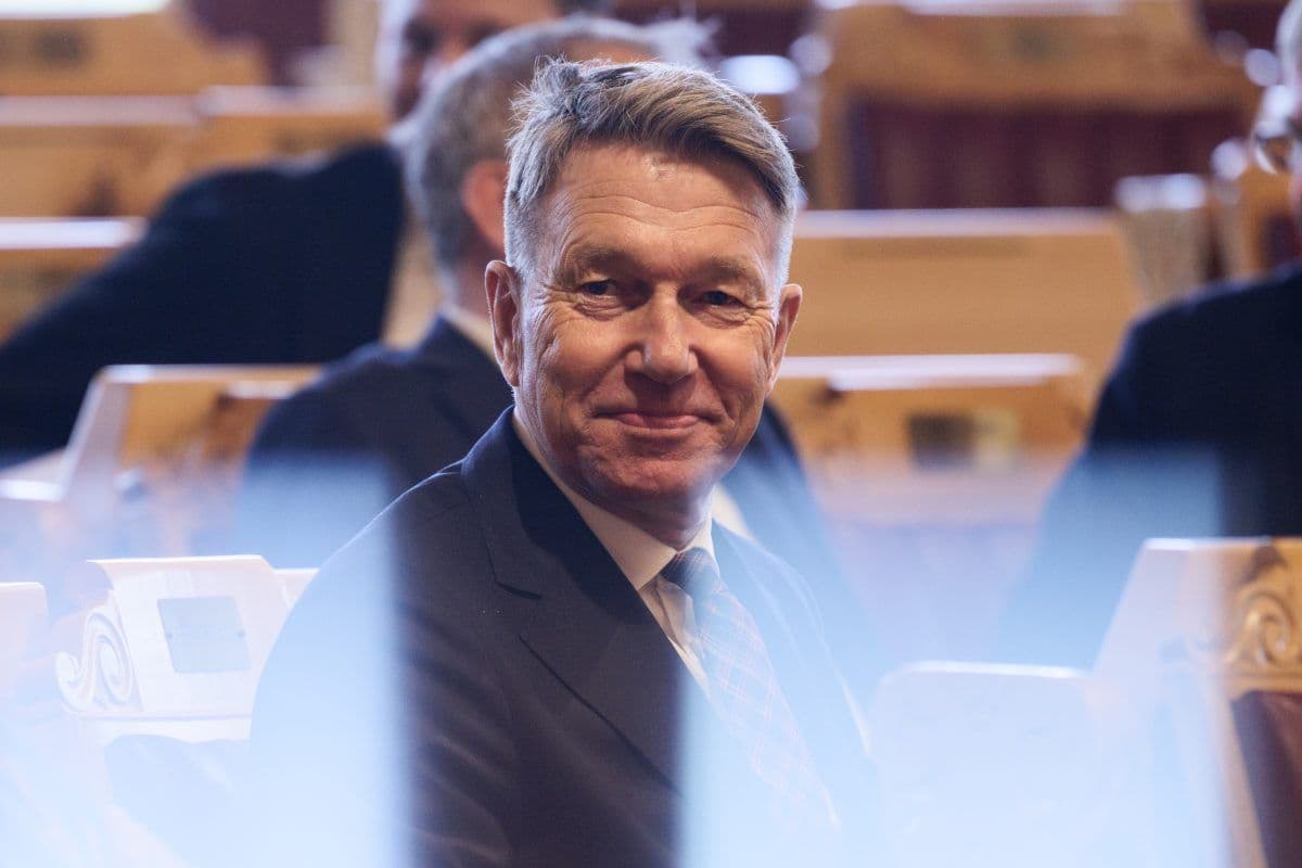 En mann i dress sitter i et auditorium av tre og smiler litt, med andre mennesker uskarpe i bakgrunnen.