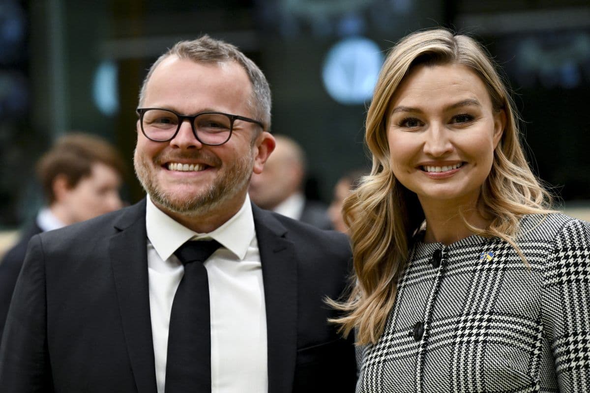 En mann i dress og slips står ved siden av en kvinne i rutete antrekk og smiler innendørs på et arrangement.