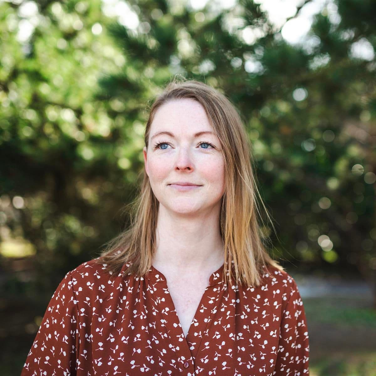 En blond kvinne med blå øyne smiler mildt, ser opp og til venstre, mot en uskarp grønn bakgrunn.