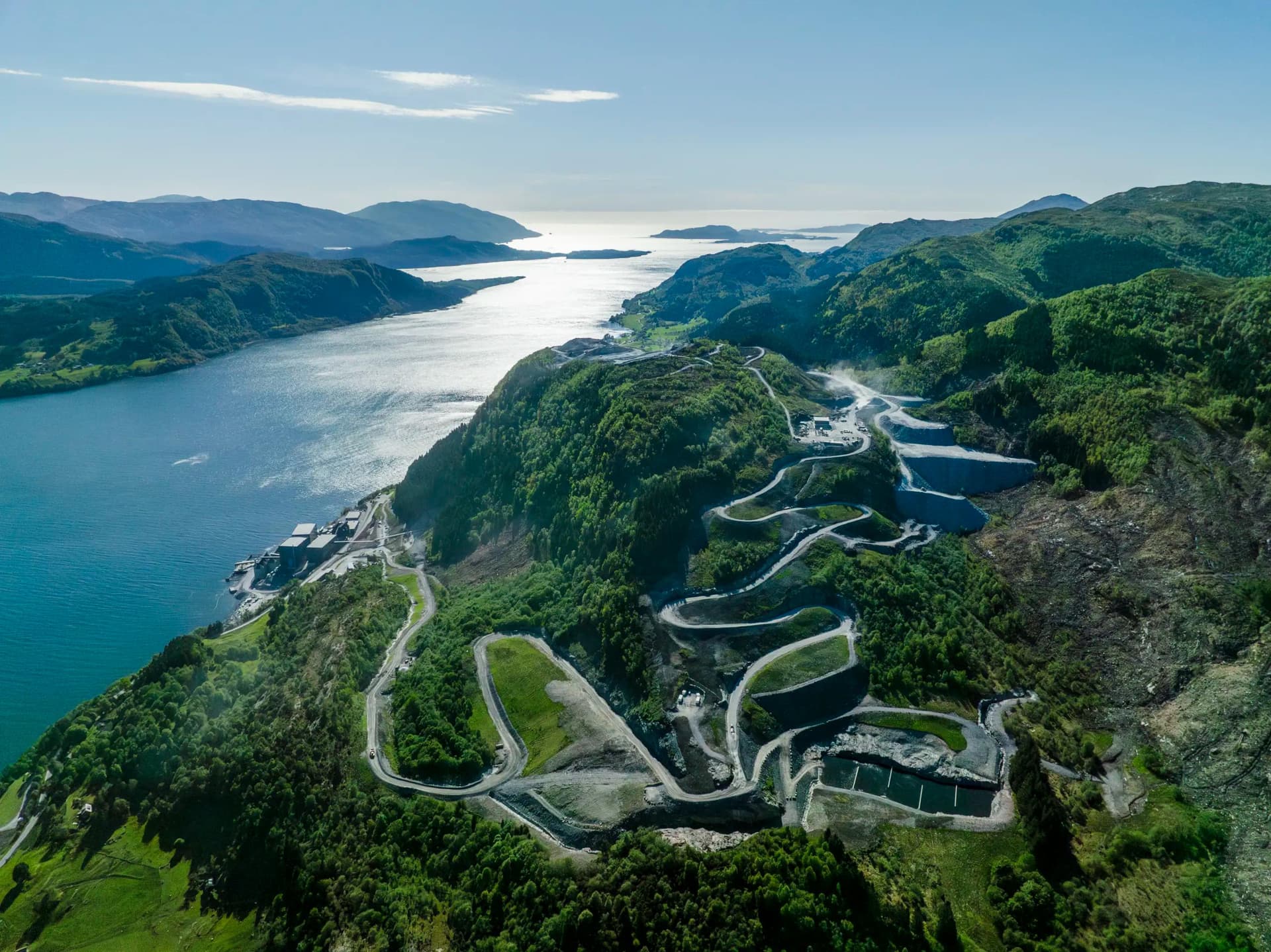 Fugleperspektiv av svingete veier og terrassert bebyggelse i en grønn åsside med utsikt over en solbelyst fjord.