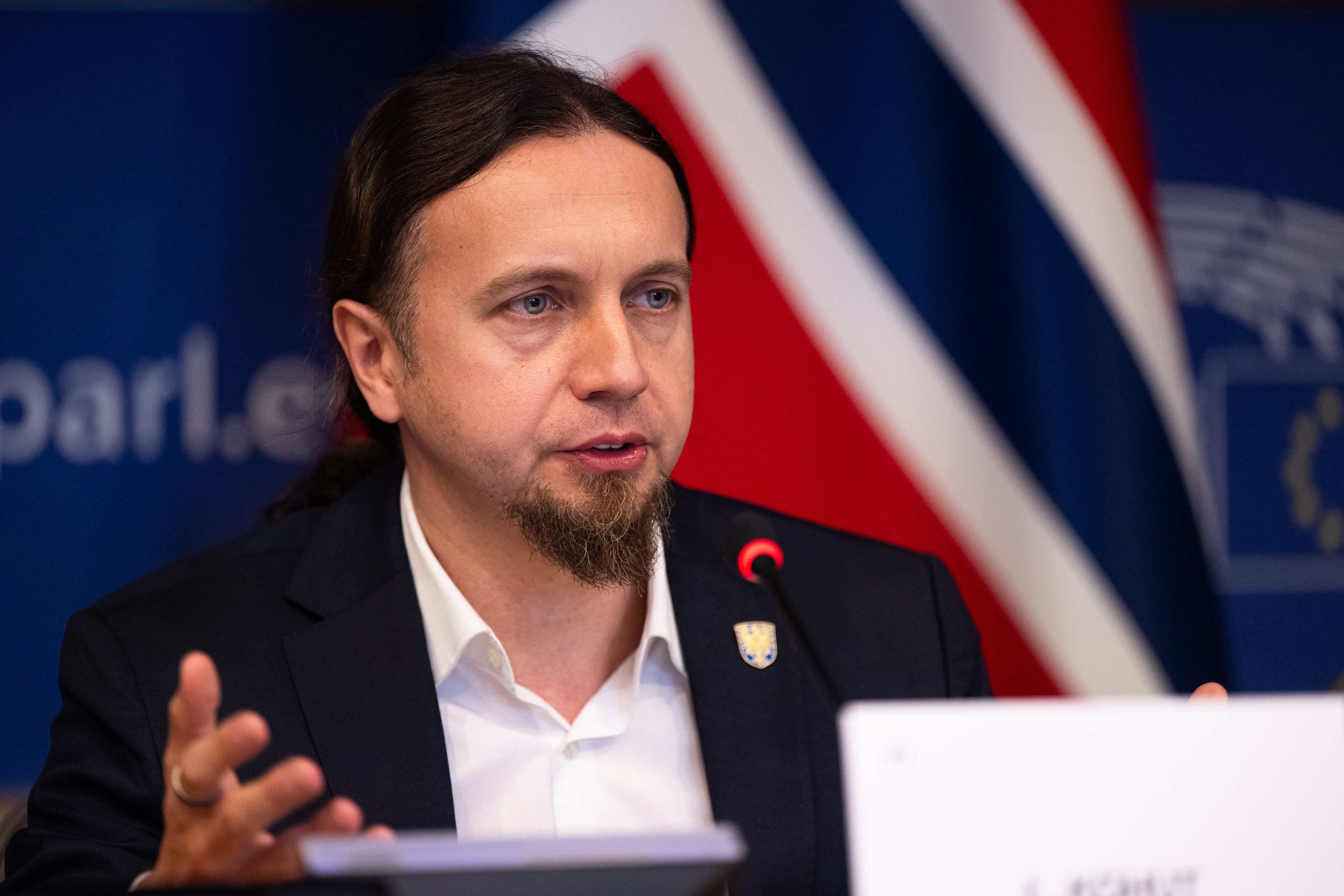 En mann med langt, mørkt hår og fippskjegg, iført dress, taler fra en talerstol, med britiske flagg og EU-flagg i bakgrunnen.