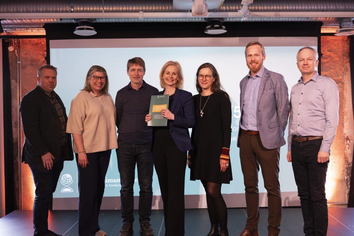 Sju personer står på en scene, en med sertifikat. En presentasjonsskjerm er i bakgrunnen.