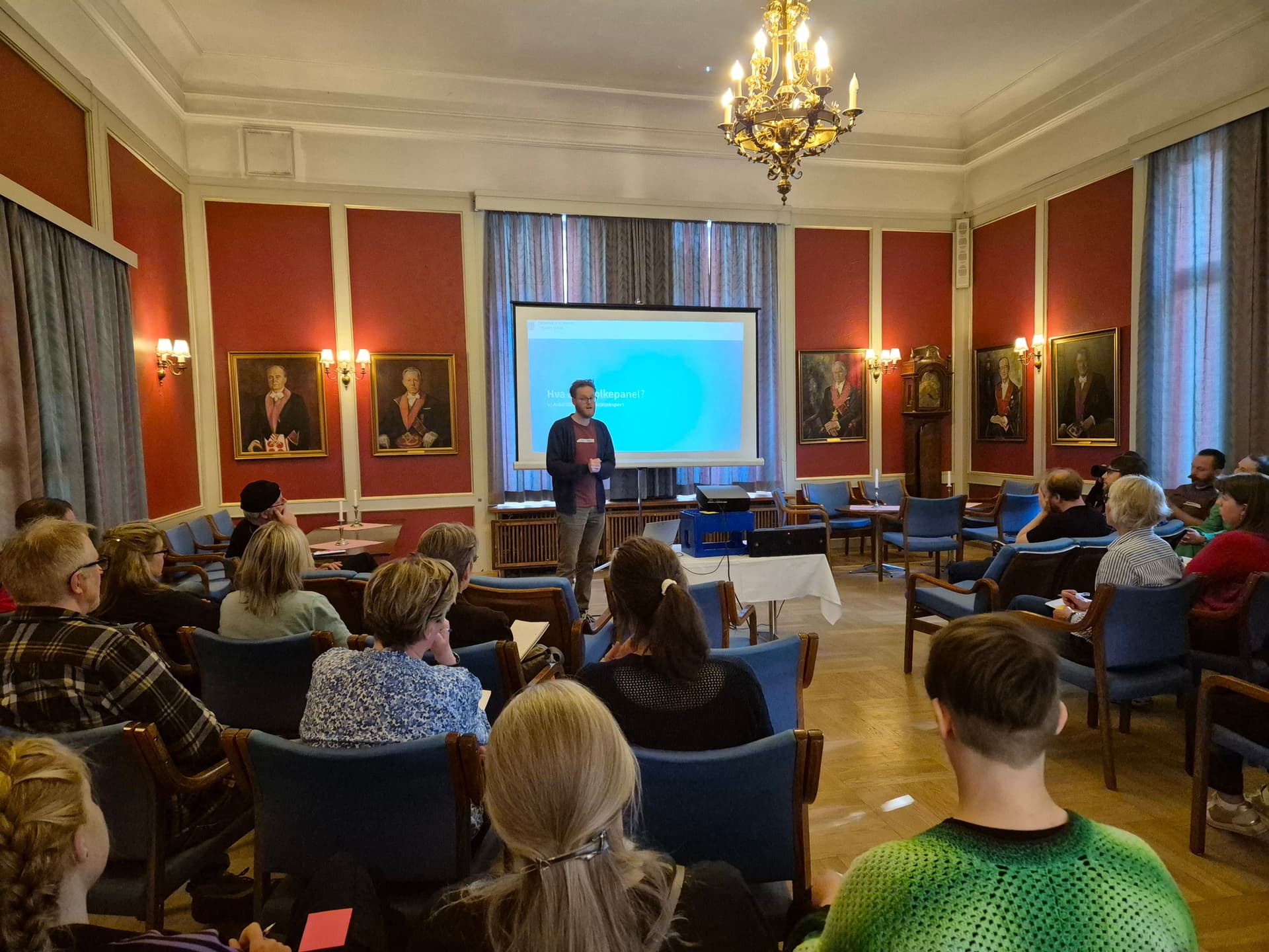 En person holder en presentasjon for et publikum i et formelt rom med røde vegger, malerier og en projektorskjerm.