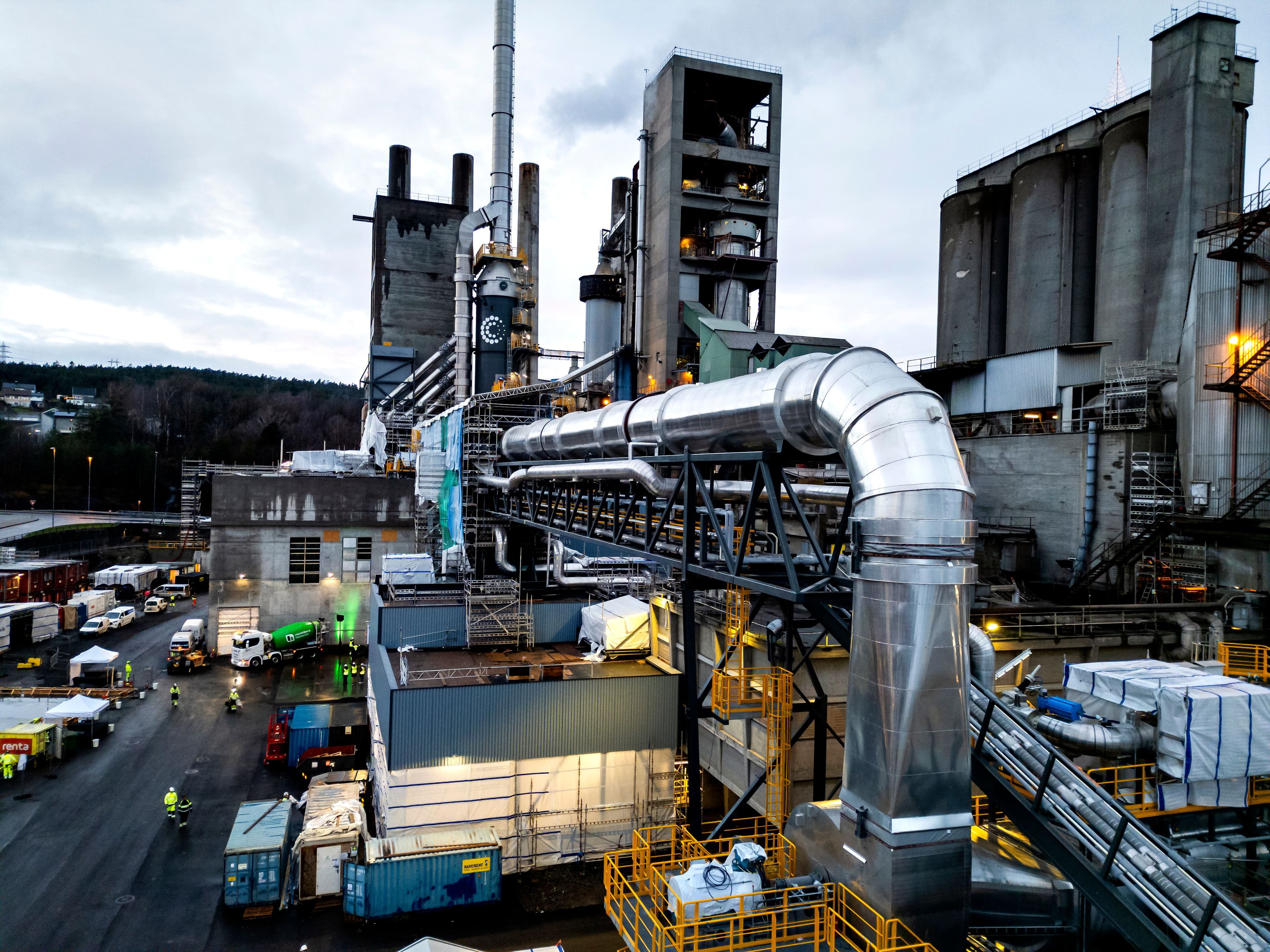 Stort industrianlegg med høye betongkonstruksjoner, siloer og skorsteiner, med omfattende rørsystemer og aktivitet på bakkeplan.