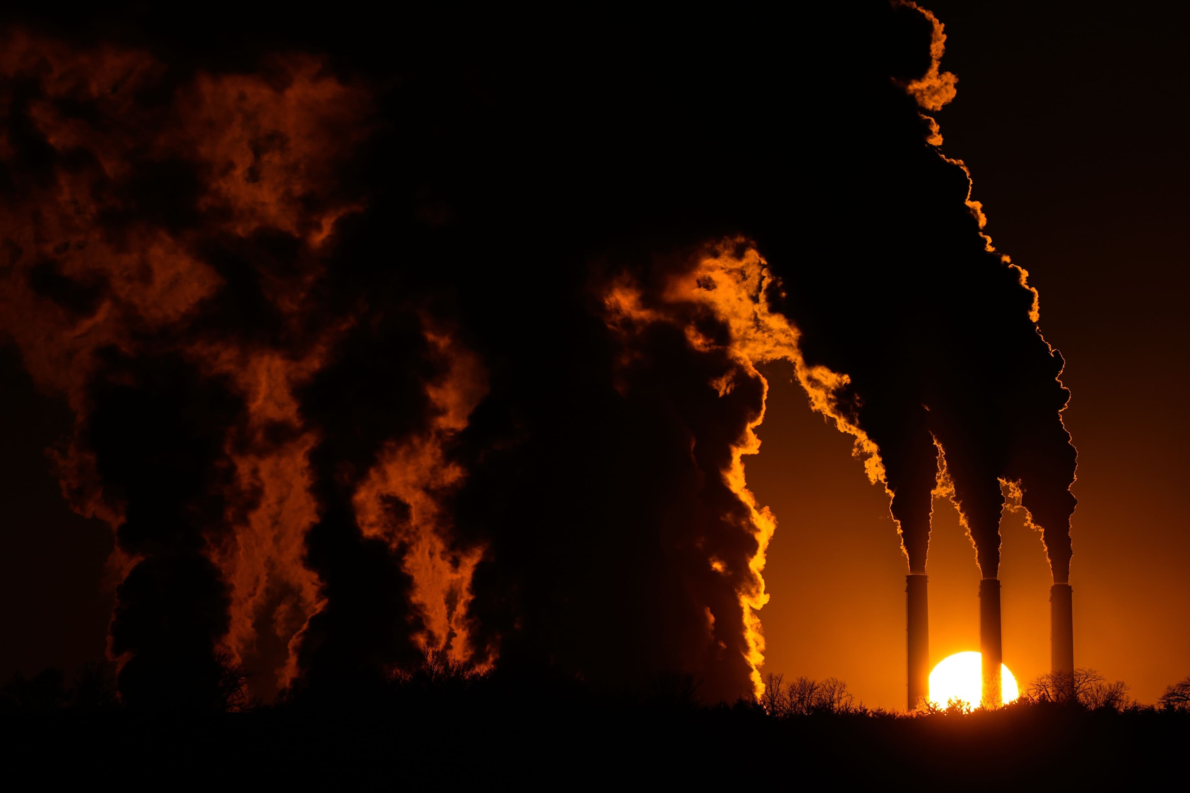 Mørk røyk velter ut av industrielle skorsteiner, opplyst bakfra av en oransje solnedgang.