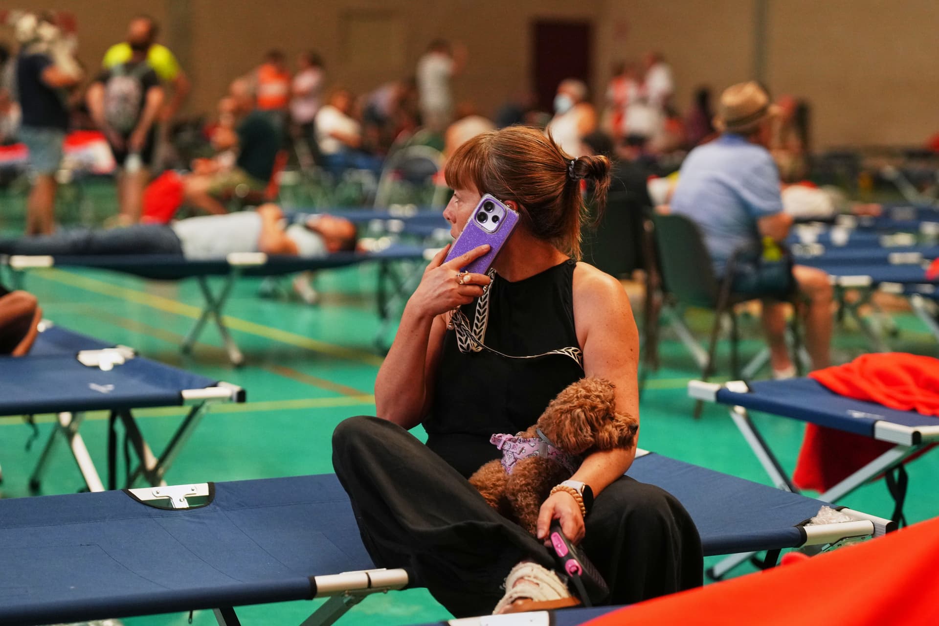 En kvinne sitter på en blå feltseng sammen med en liten brun hund og snakker i en lilla telefon i et overfylt nødhus.