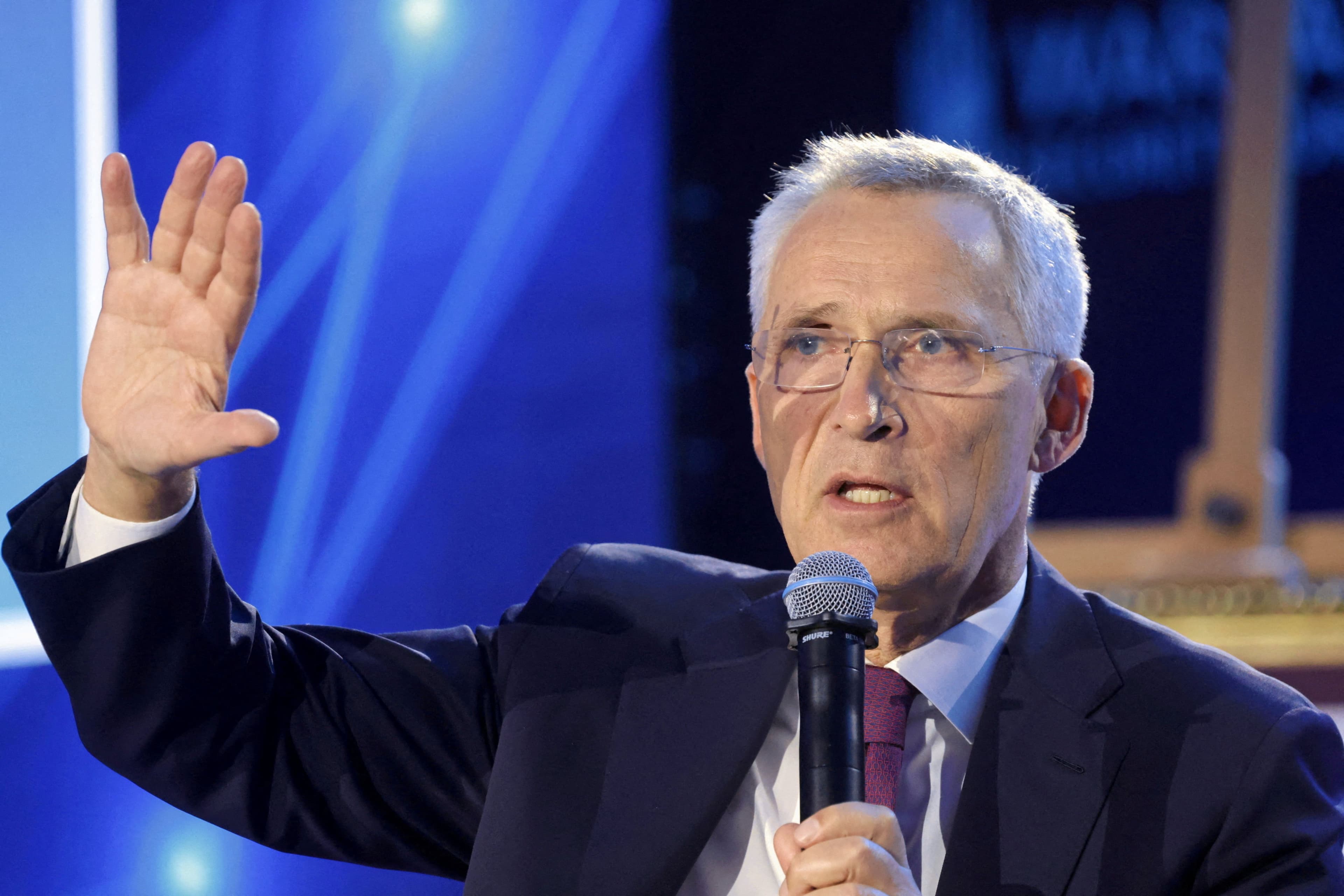 En mann med grått hår og briller snakker i en mikrofon, med venstre hånd løftet i en gest.