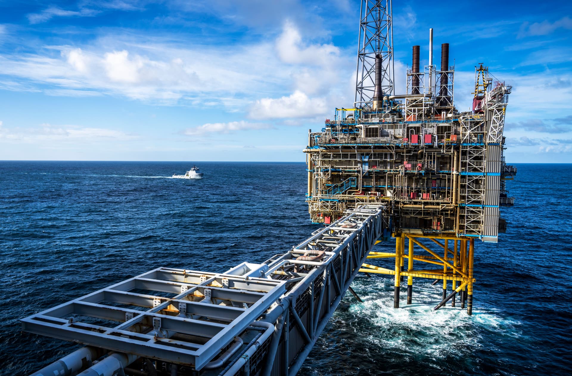 En offshore oljeplattform forbundet med en gangbro over det dype blå havet, med en båt i det fjerne under en delvis skyet himmel.