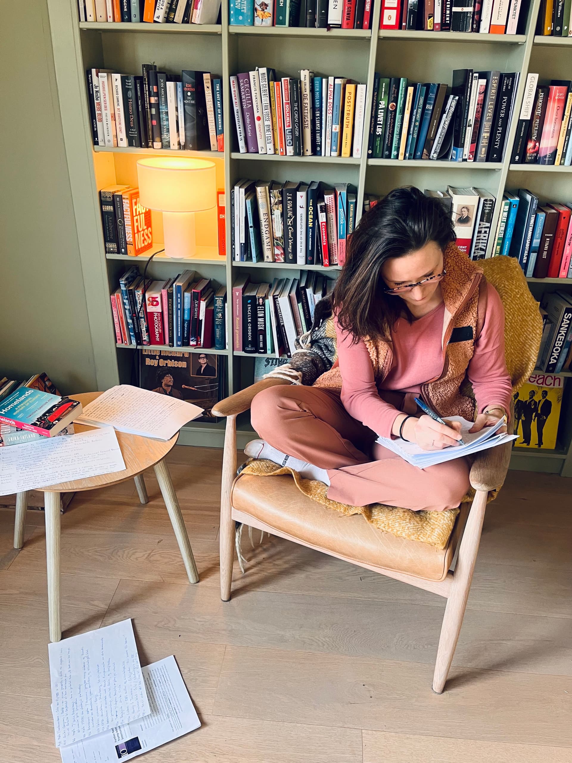 A person sits cross-legged in an armchair, writing in a notebook, surrounded by bookshelves and papers.