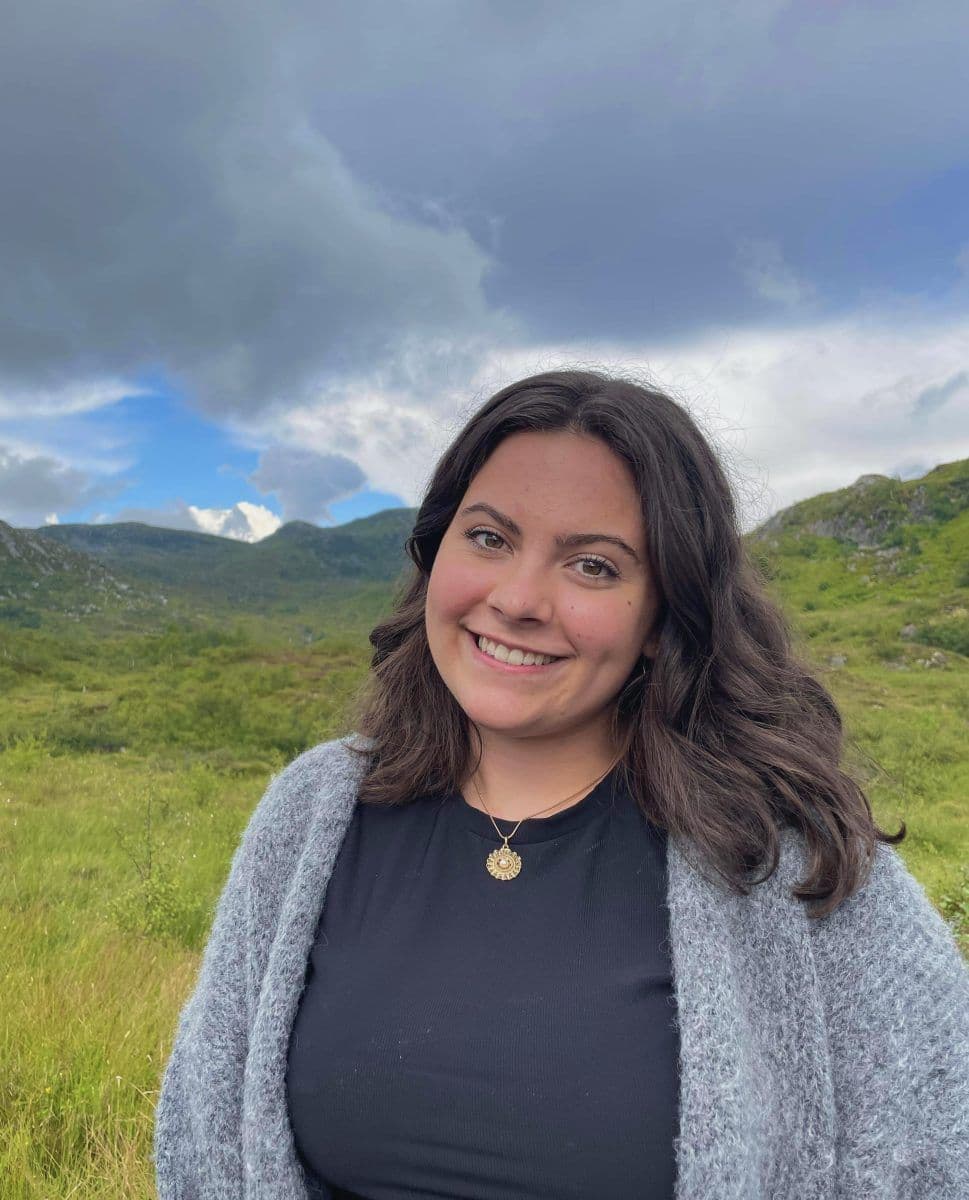 En person som smiler utendørs, står i en gressmark med åser og en overskyet himmel i bakgrunnen.