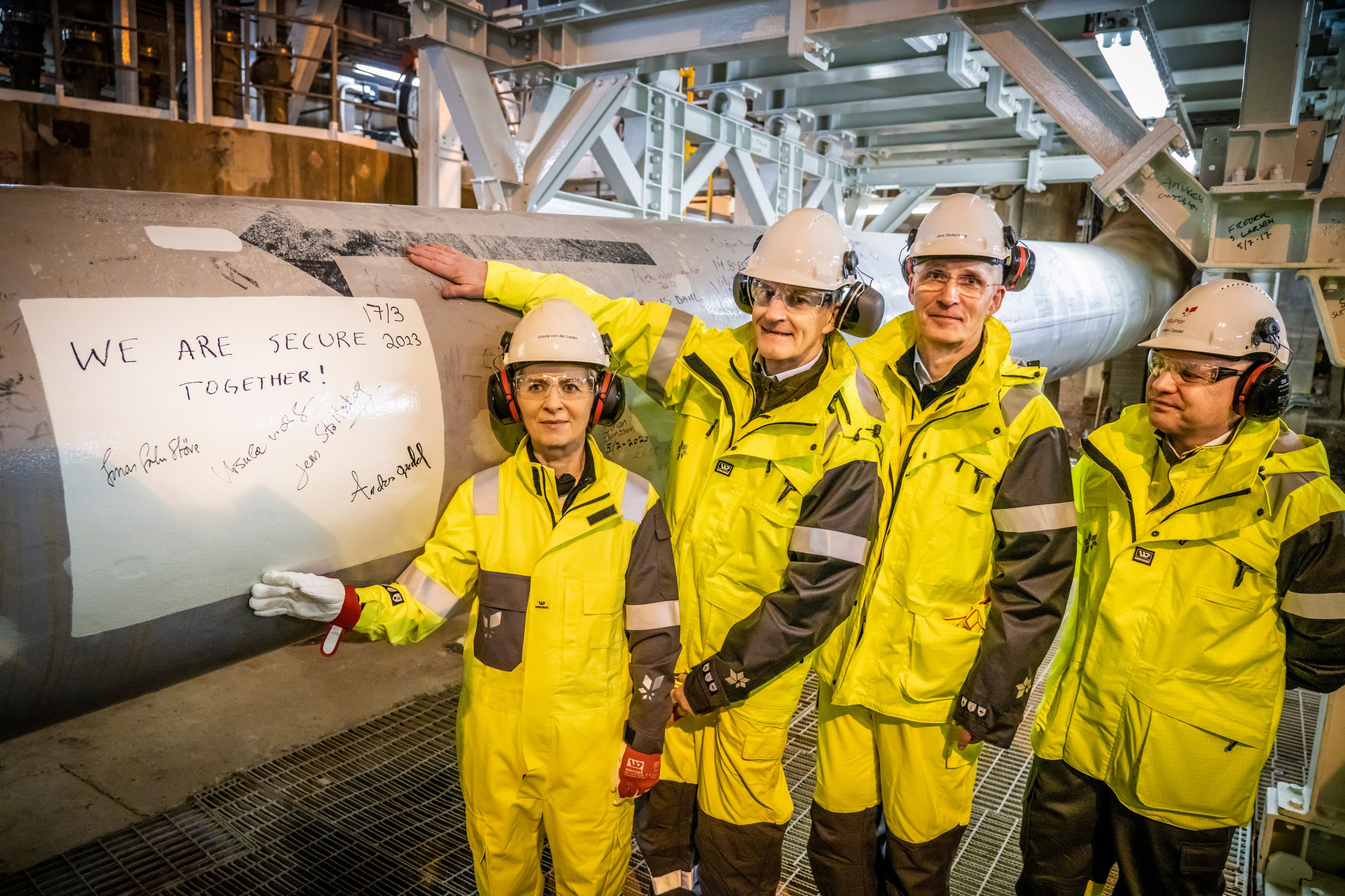 Fire personer i verneutstyr ved siden av et stort industrirør, der et ark med teksten «VI ER TRYGGERE SAMMEN!» med signaturer og datoen 2017 er festet.