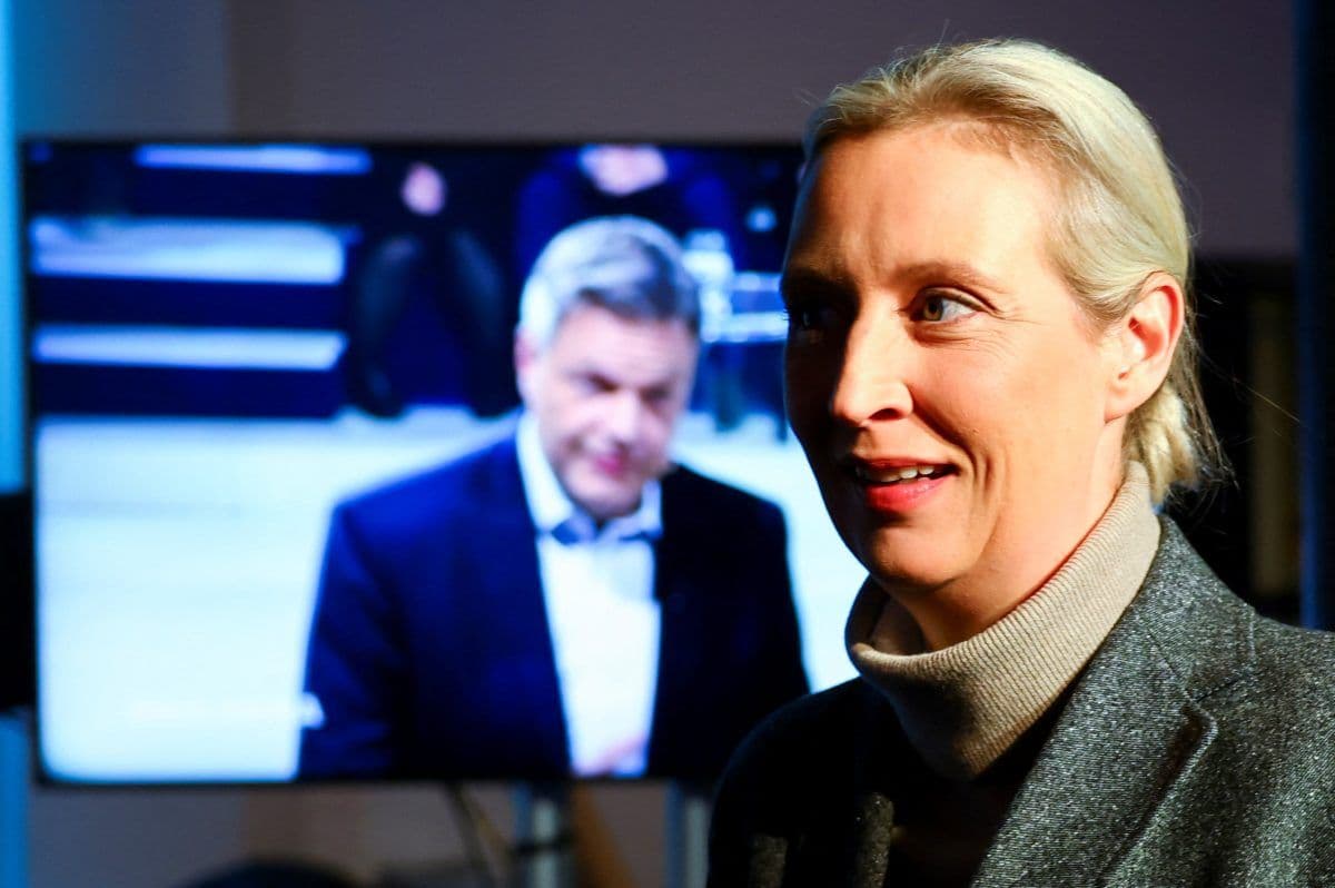 En kvinne i fokus står i forgrunnen med en uskarp mann på en TV-skjerm bak seg. Hun har på seg en grå frakk og ser ut til å være i studio eller kringkasting.