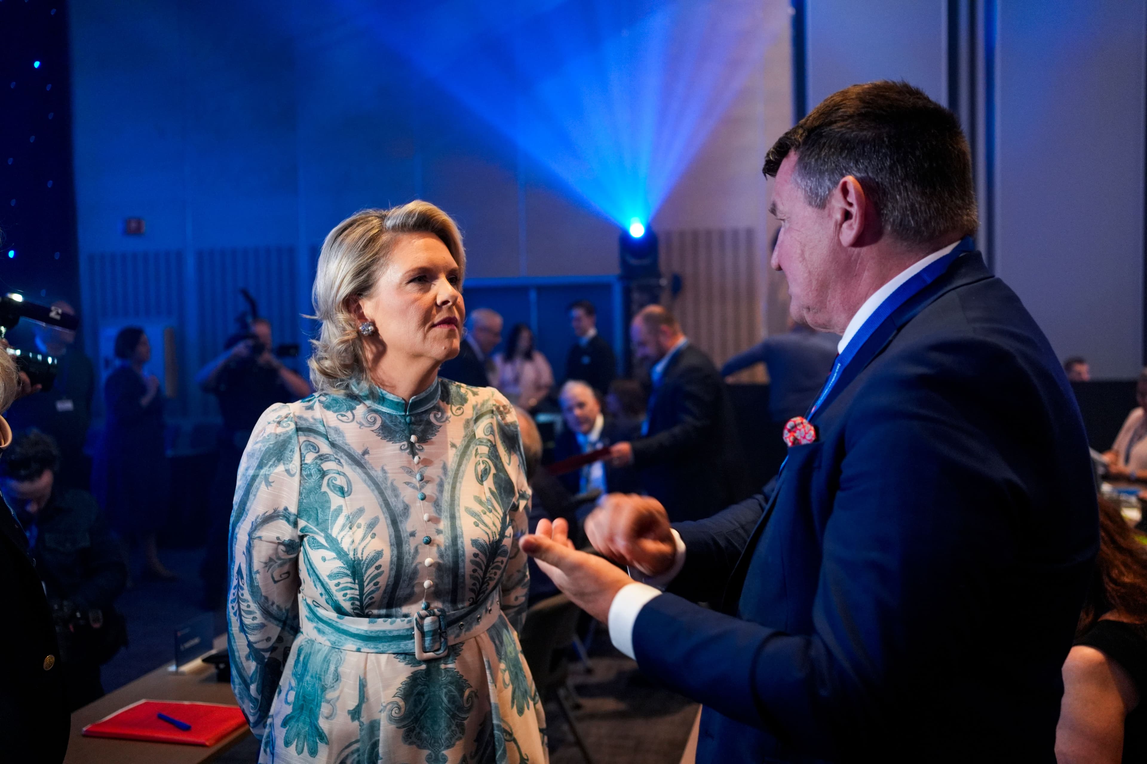 A woman in a patterned dress looks at a man in a suit who is gesturing with his hands in a blue-lit event space.