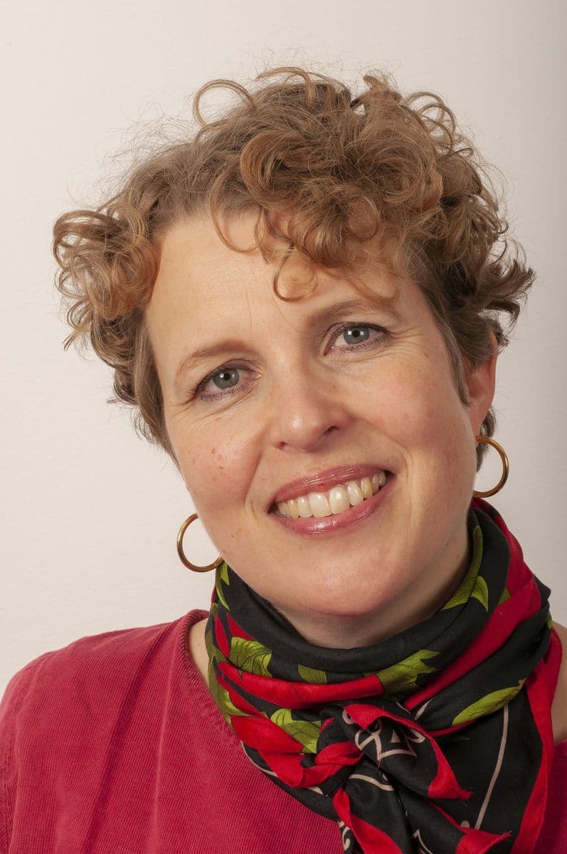 En kvinne med kort, krøllete hår, iført bøyleøreringer, en rød topp og et svart og rødt mønstret skjerf, smiler til kameraet mot en enkel bakgrunn.