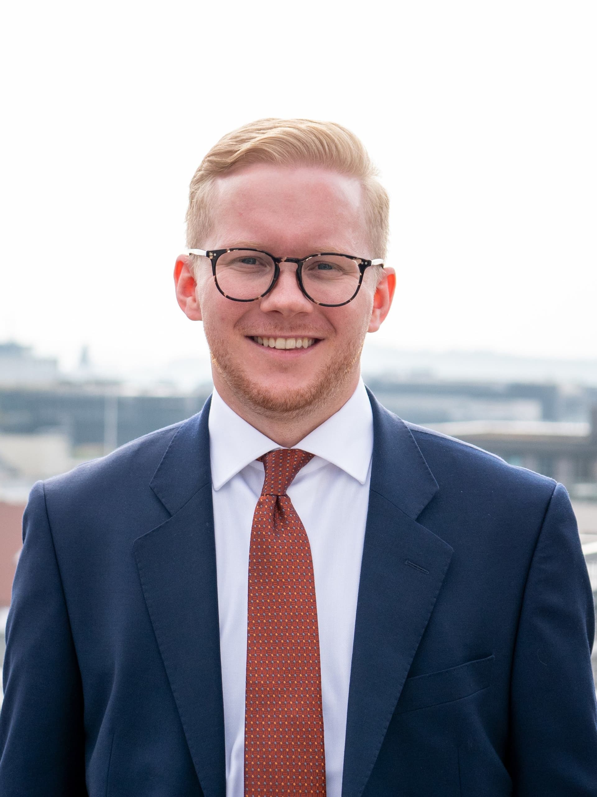 En smilende ung mann med blondt hår og briller i blå dress og mønstret oransje slips, utendørs.
