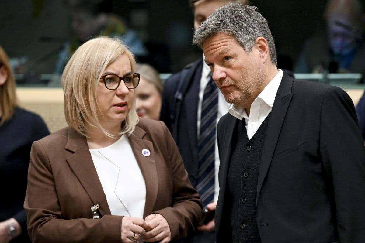 To personer i formell antrekk er engasjert i samtale i en profesjonell setting, omgitt av andre i bakgrunnen.