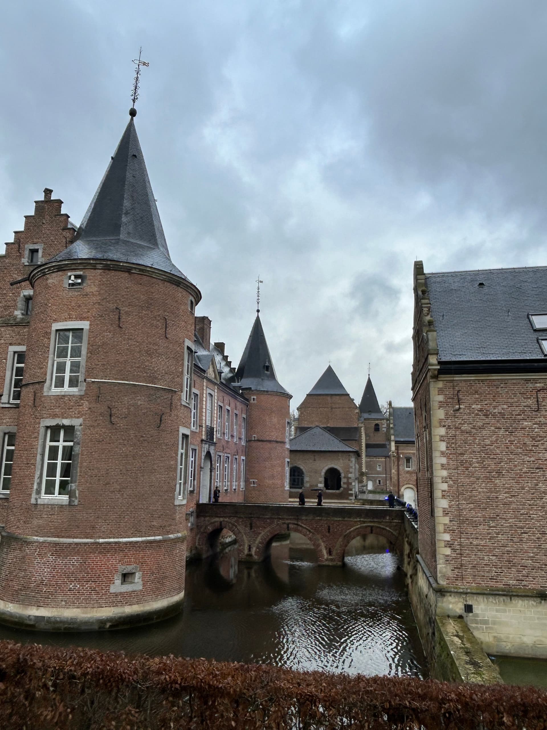 Et mursteinslott med et rundt tårn og en bro over en vollgrav under en overskyet himmel.