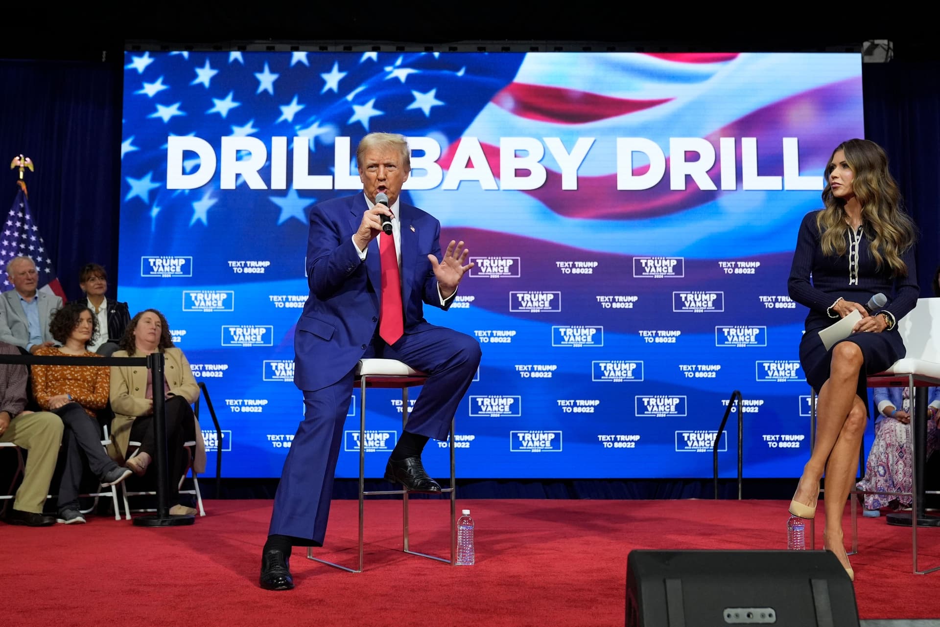 Donald Trump speaks into a microphone while seated on a stool next to a woman, with a large screen behind them displaying "DRILL BABY DRILL" on an American flag.