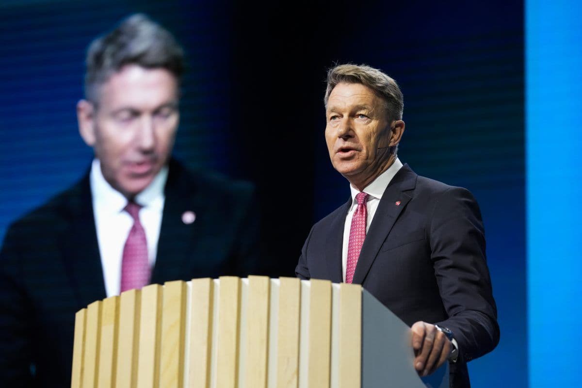 En mann i dress og rødt slips taler på et podium, mens bildet av ham vises på en stor skjerm bak ham.