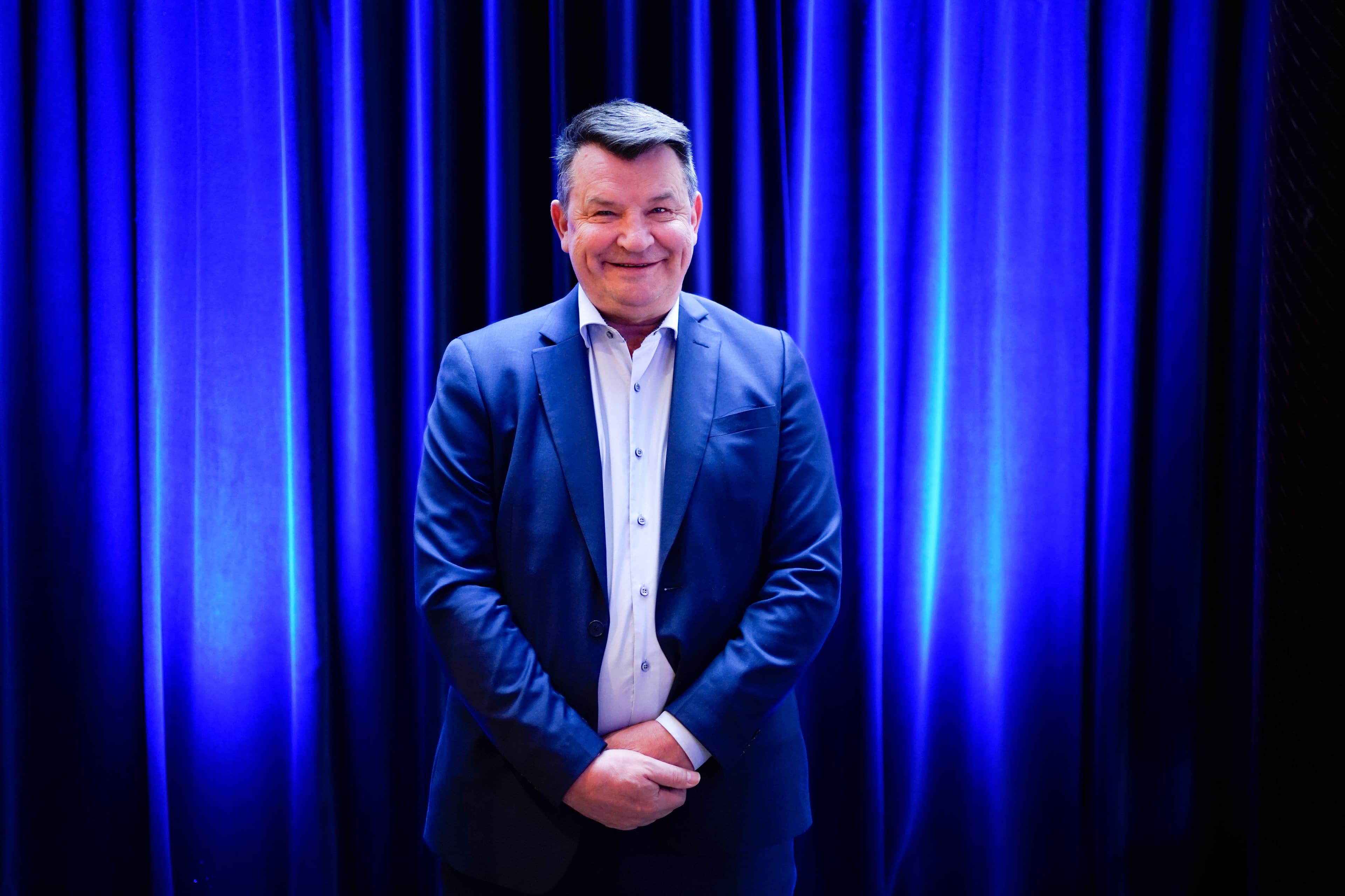 En smilende mann i blå dress står foran et blått sceneteppe.