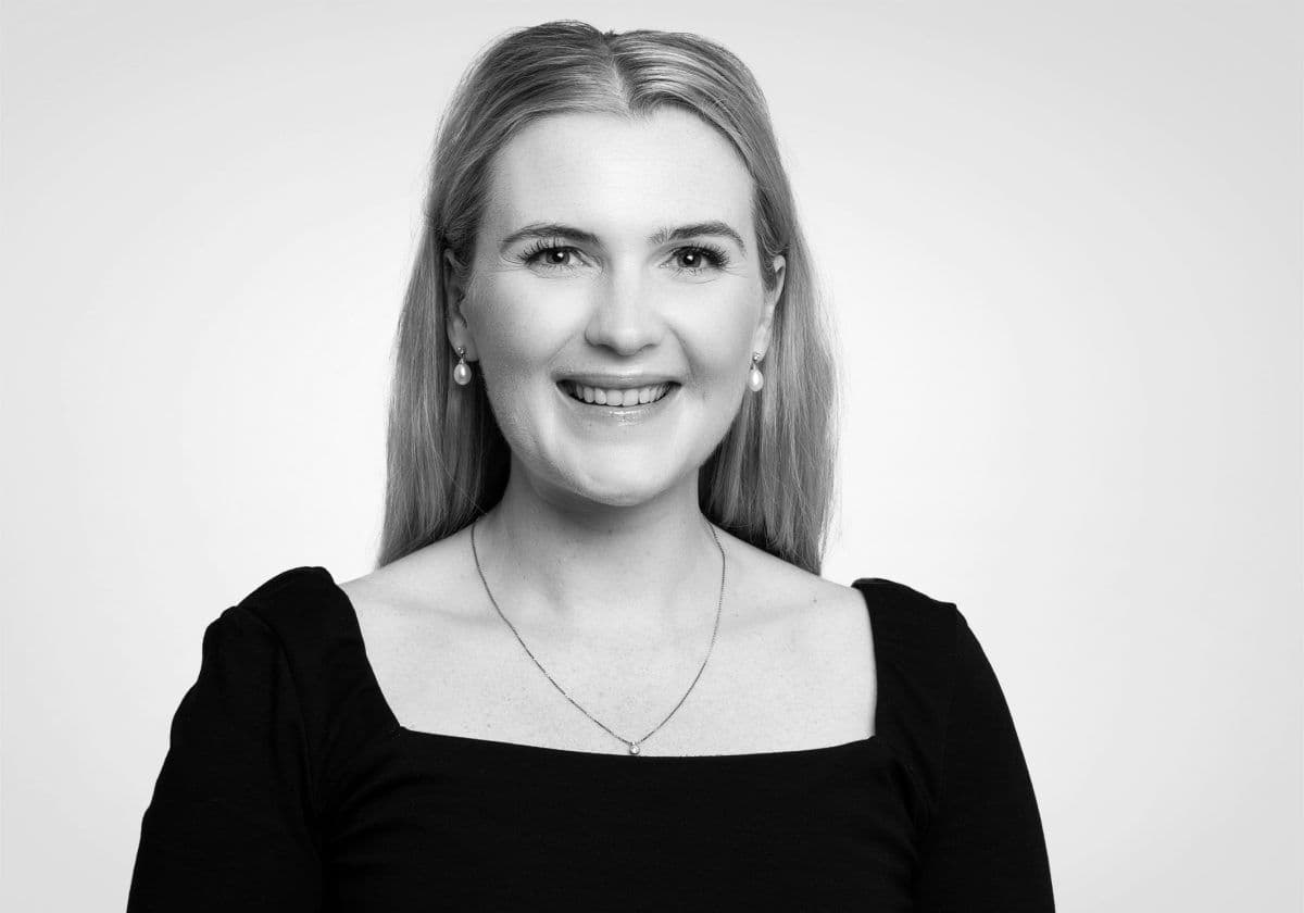 Portrett av en kvinne med langt lyst hår, iført en svart topp, et halskjede og øredobber, smilende til kameraet mot en vanlig lys bakgrunn.