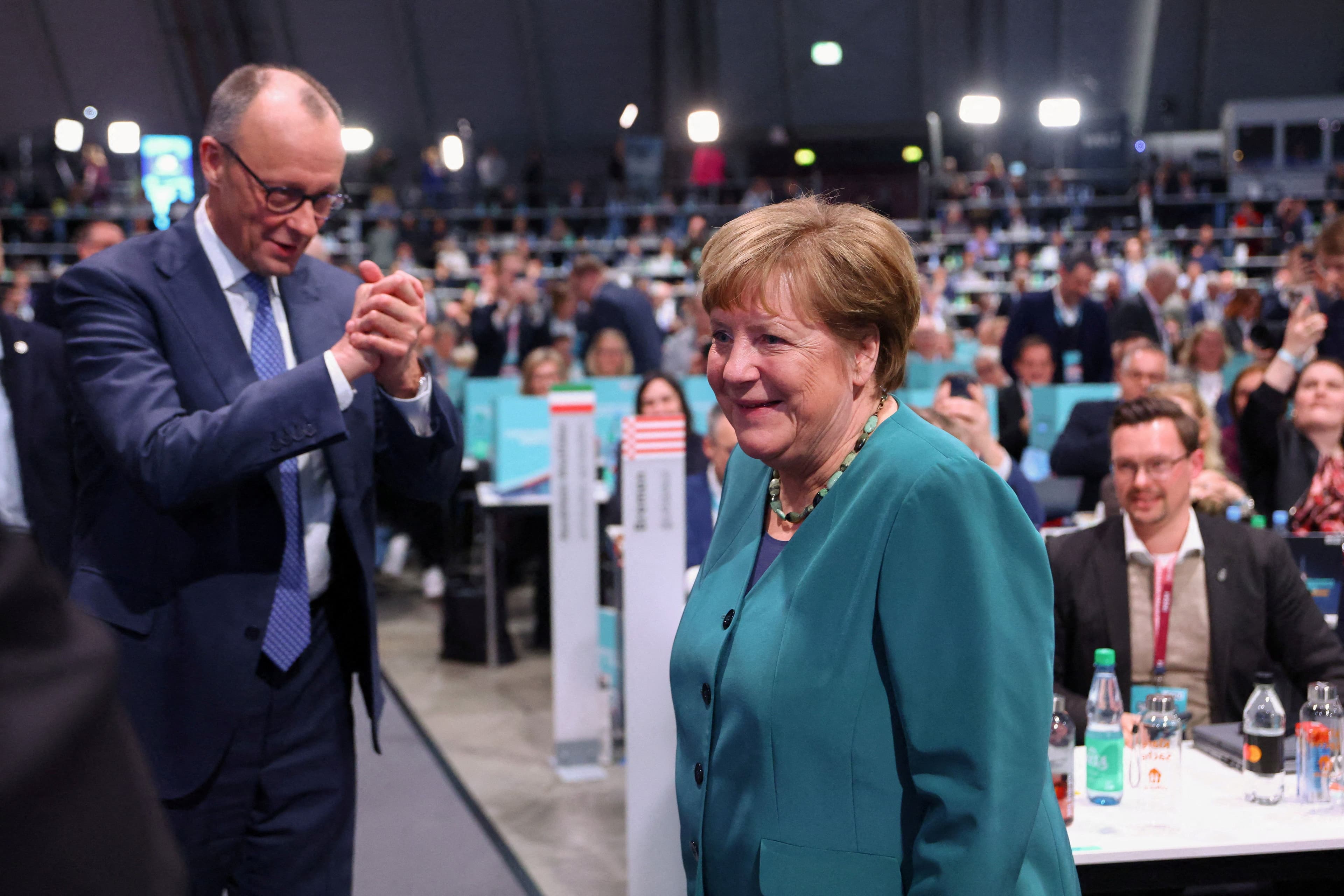Angela Merkel smiler i en turkis jakke mens Friedrich Merz applauderer henne på et arrangement.