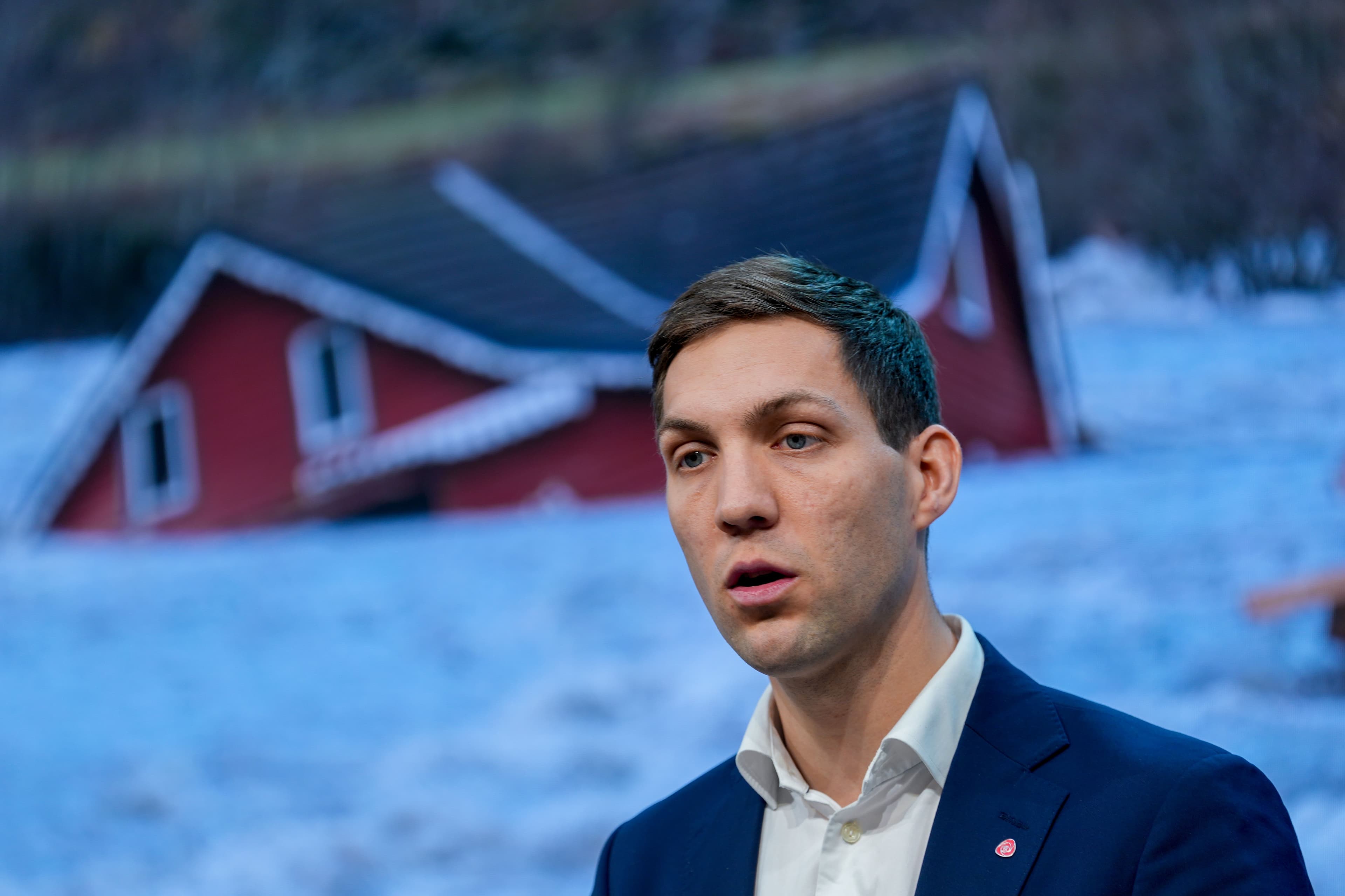 En mann i dress snakker foran en uskarp bakgrunn som viser et rødt hus delvis begravd i snø.