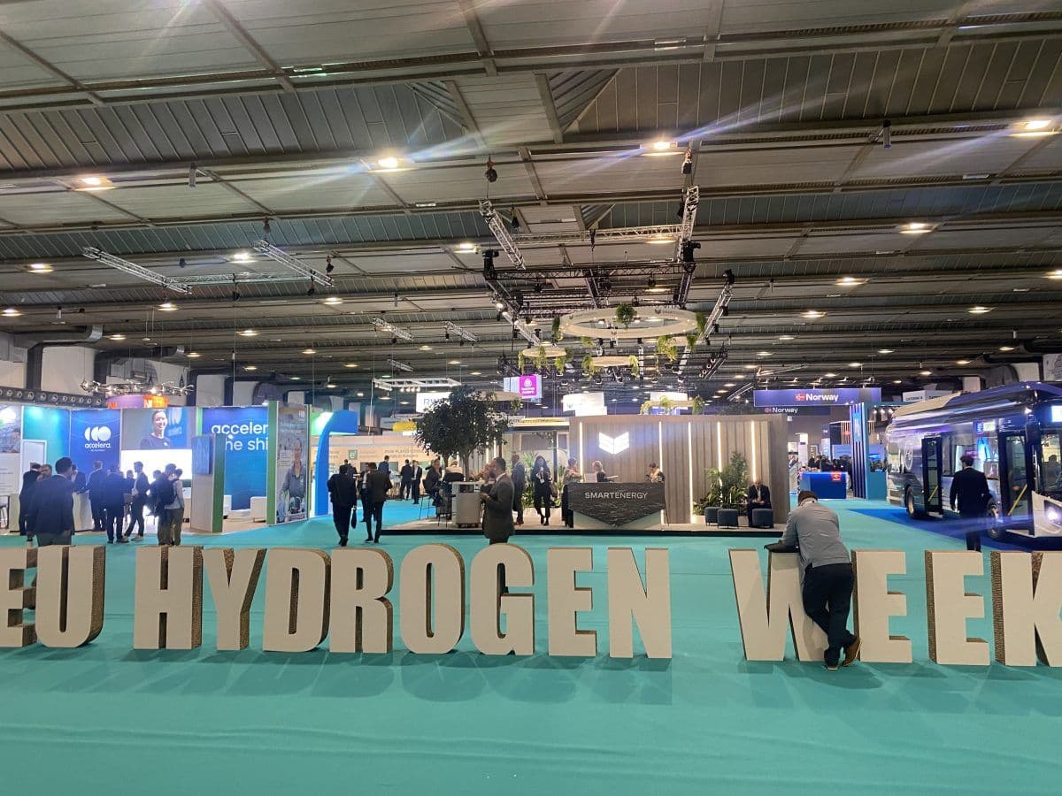 Interiør i en stor arrangementshall med "EU Hydrogen Week"-skilt, utstillingsstander og besøkende som går.