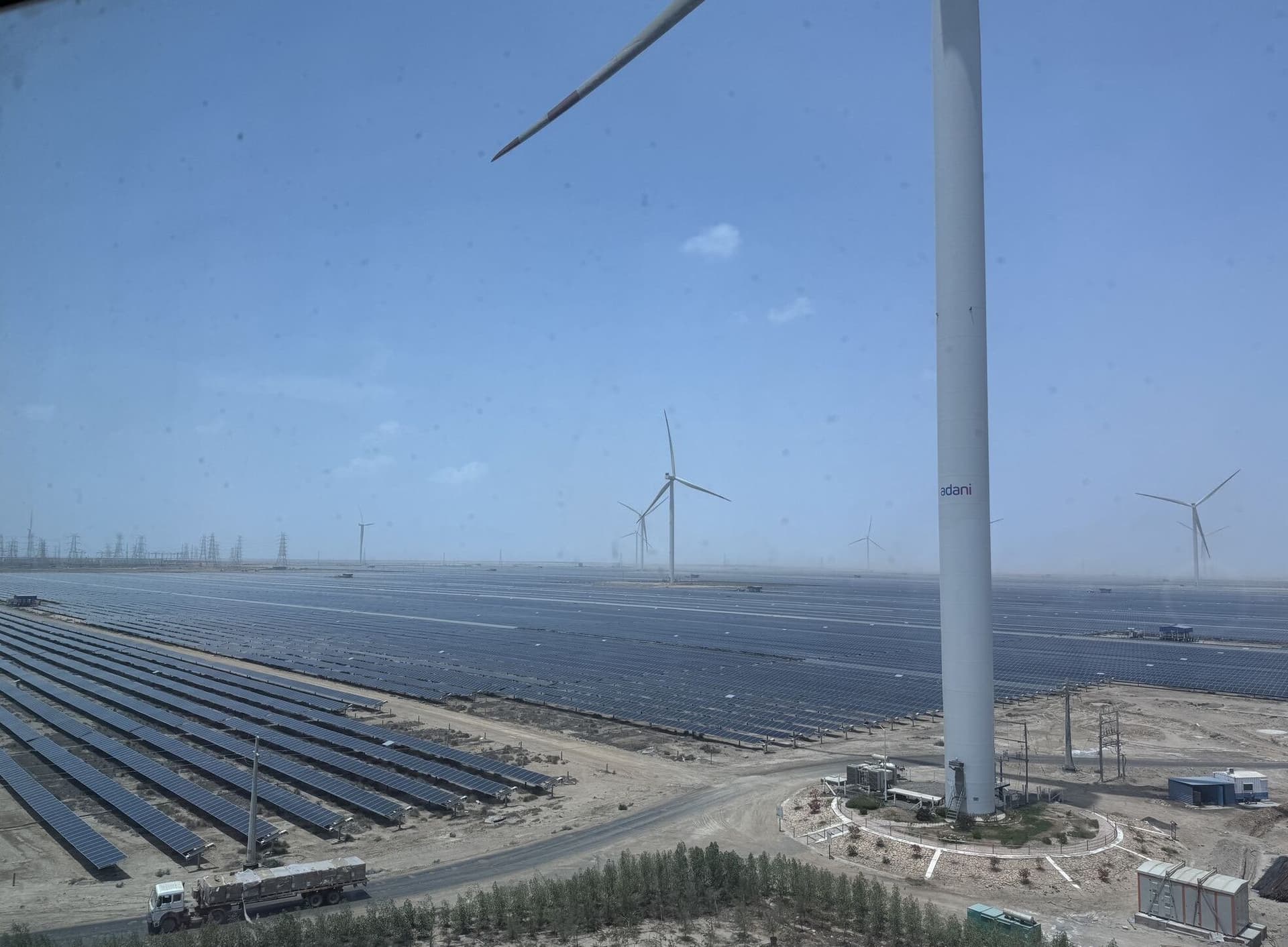 Et enormt solkraftanlegg med rekker av solcellepaneler og flere vindturbiner, inkludert én med Adani-logoen, i et tørt og karrig landskap.