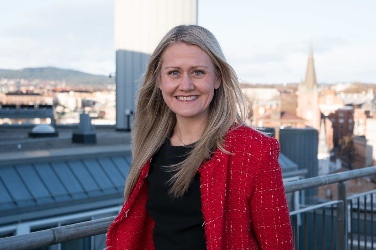 En kvinne med langt blondt hår, iført rød jakke og svart topp, står på et hustak med et bylandskap i bakgrunnen.