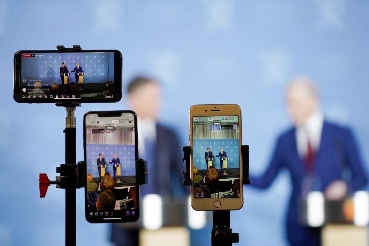 Telefoner montert på stativ tar opp to personer som snakker på en pressekonferanse, sett mot blå bakgrunn.