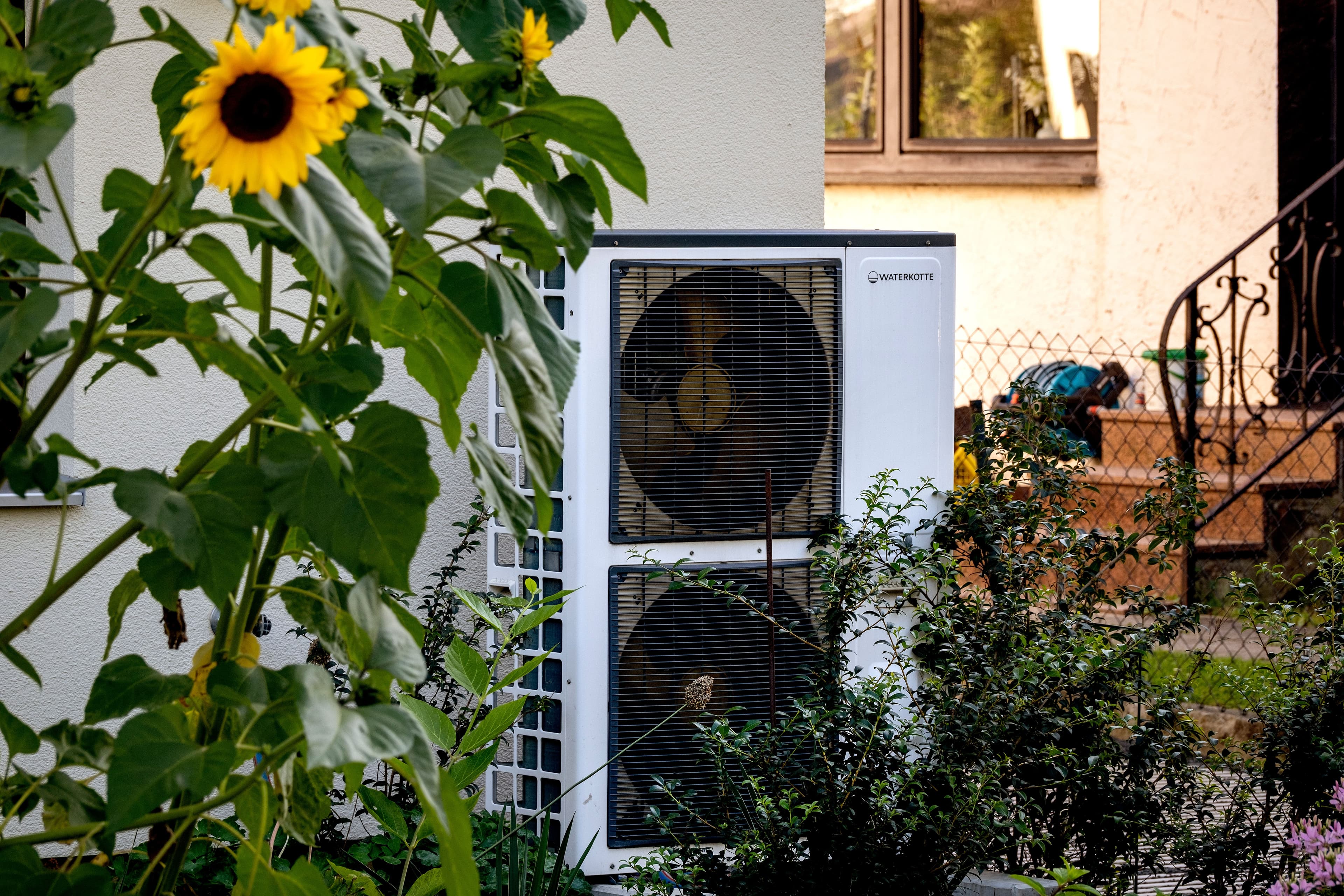 A white outdoor heat pump unit with two fans, partially obscured by green plants and a sunflower.